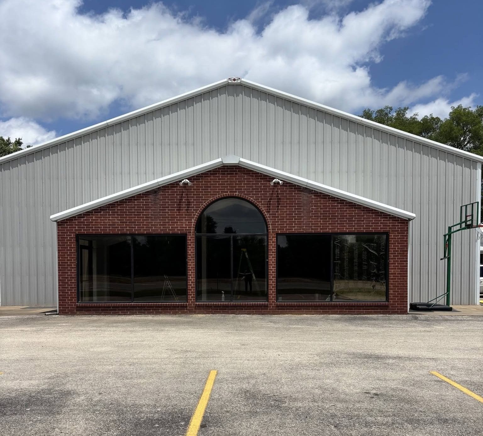 Front view of a red-brick and white metal building with an arched entrance and parking lot lines in front