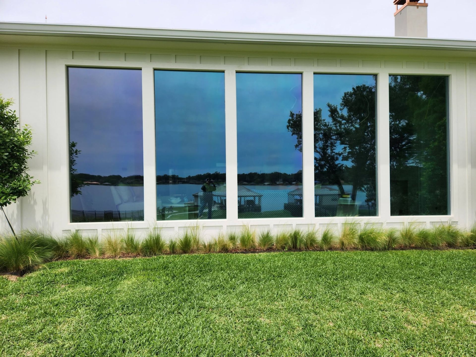 White house exterior with large windows reflecting a lake and trees, above a green lawn.