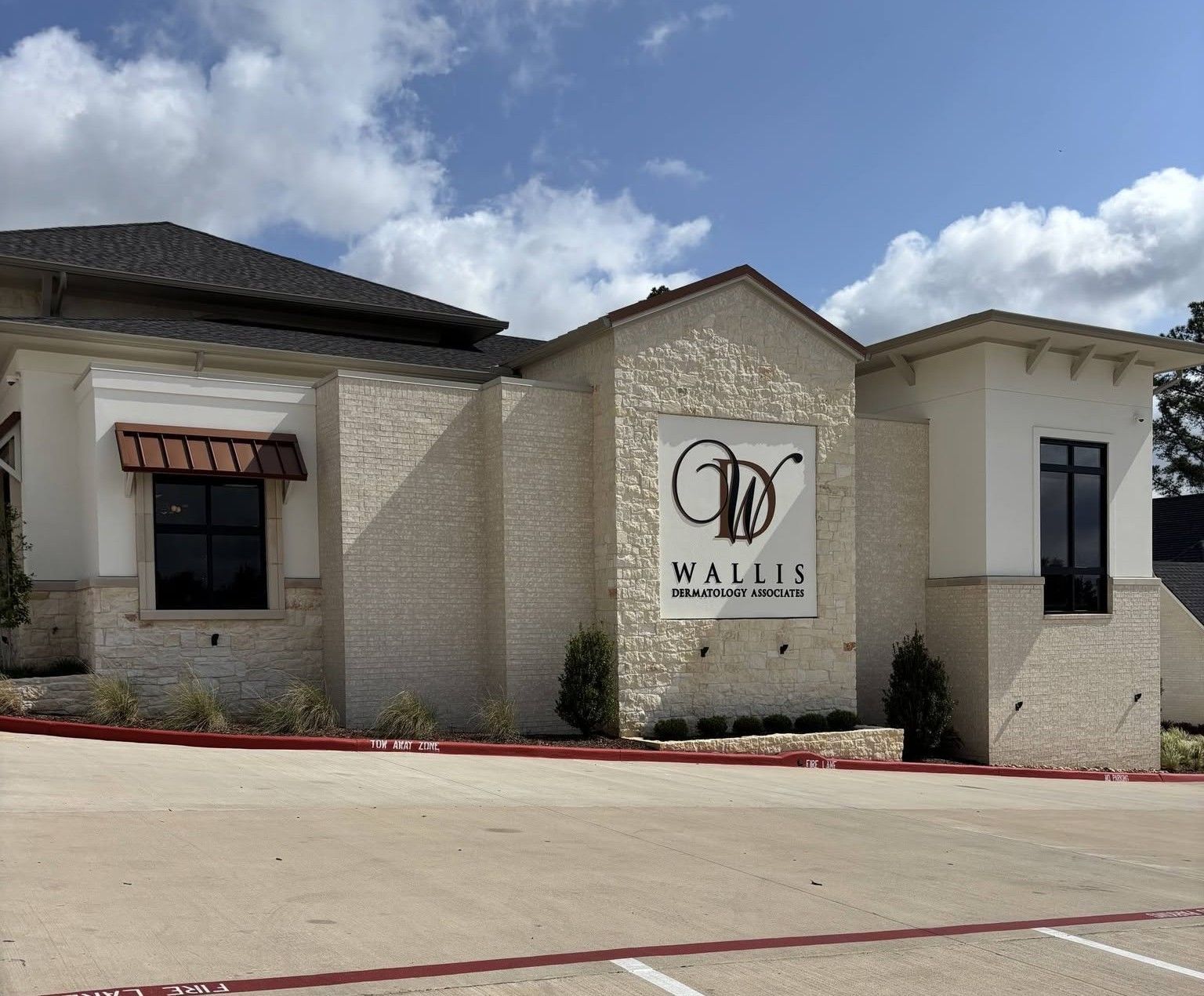 Exterior of Wallis building with stone facade, black roof, and large logo sign under a partly cloudy sky