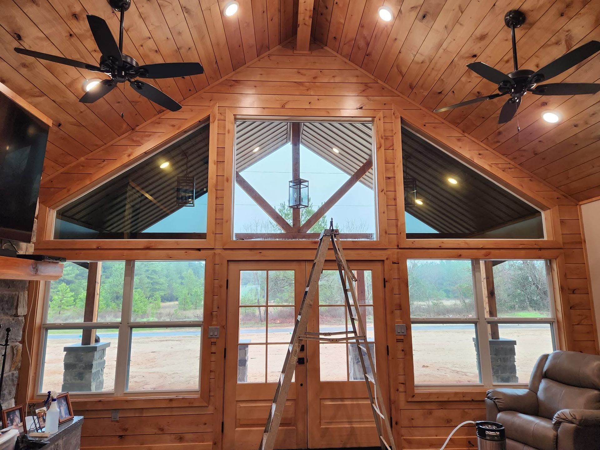 Wood-paneled cabin interior with vaulted ceiling, ceiling fans, ladder, large windows, and a chair by the wall