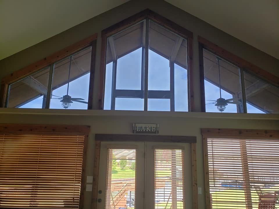 Interior view of a tall triangular window above wooden blinds and a glass door with greenery outside
