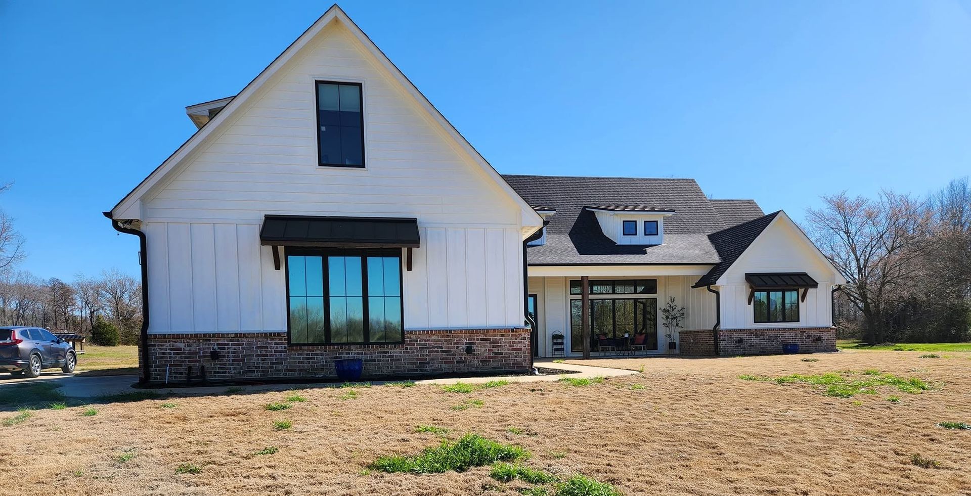 White modern farmhouse with large windows and stone base on a grassy lot under a clear blue sky