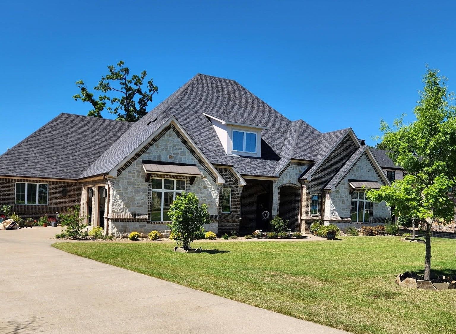 Large stone suburban house with steep gray roofs, front lawn, and a driveway under a clear blue sky