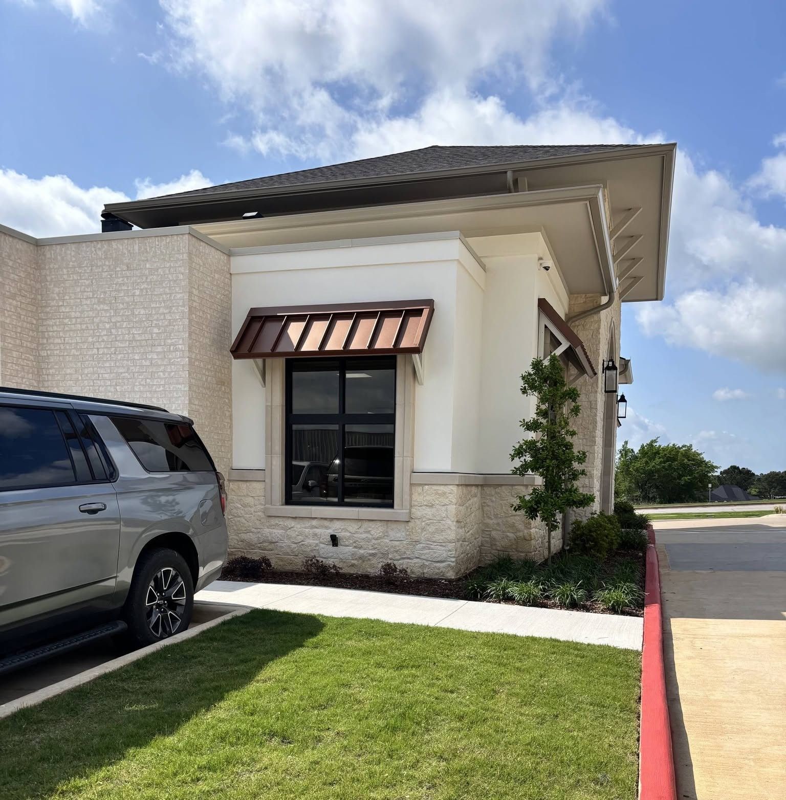 Storefront corner with small awning, grass lawn, parked SUV, and red curb under a partly cloudy sky