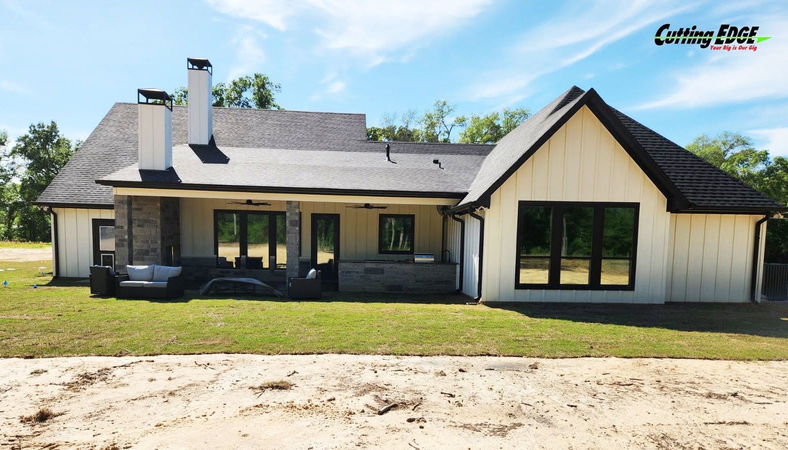 Single-story house with stone and white siding, dark roof, large windows, and a front patio under trees