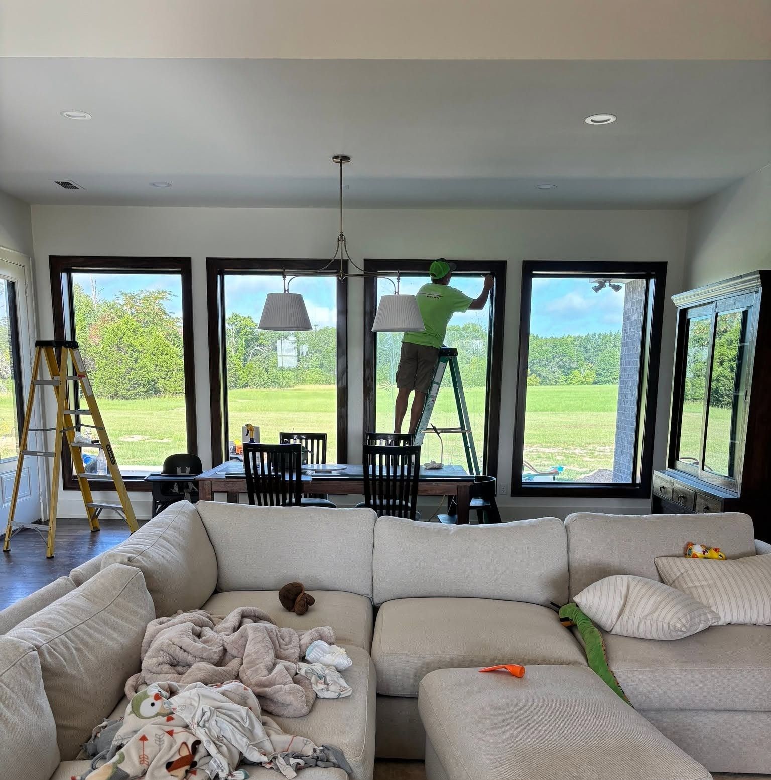 Bright living room with large windows, two ladders, and people hanging a green figure on the wall.