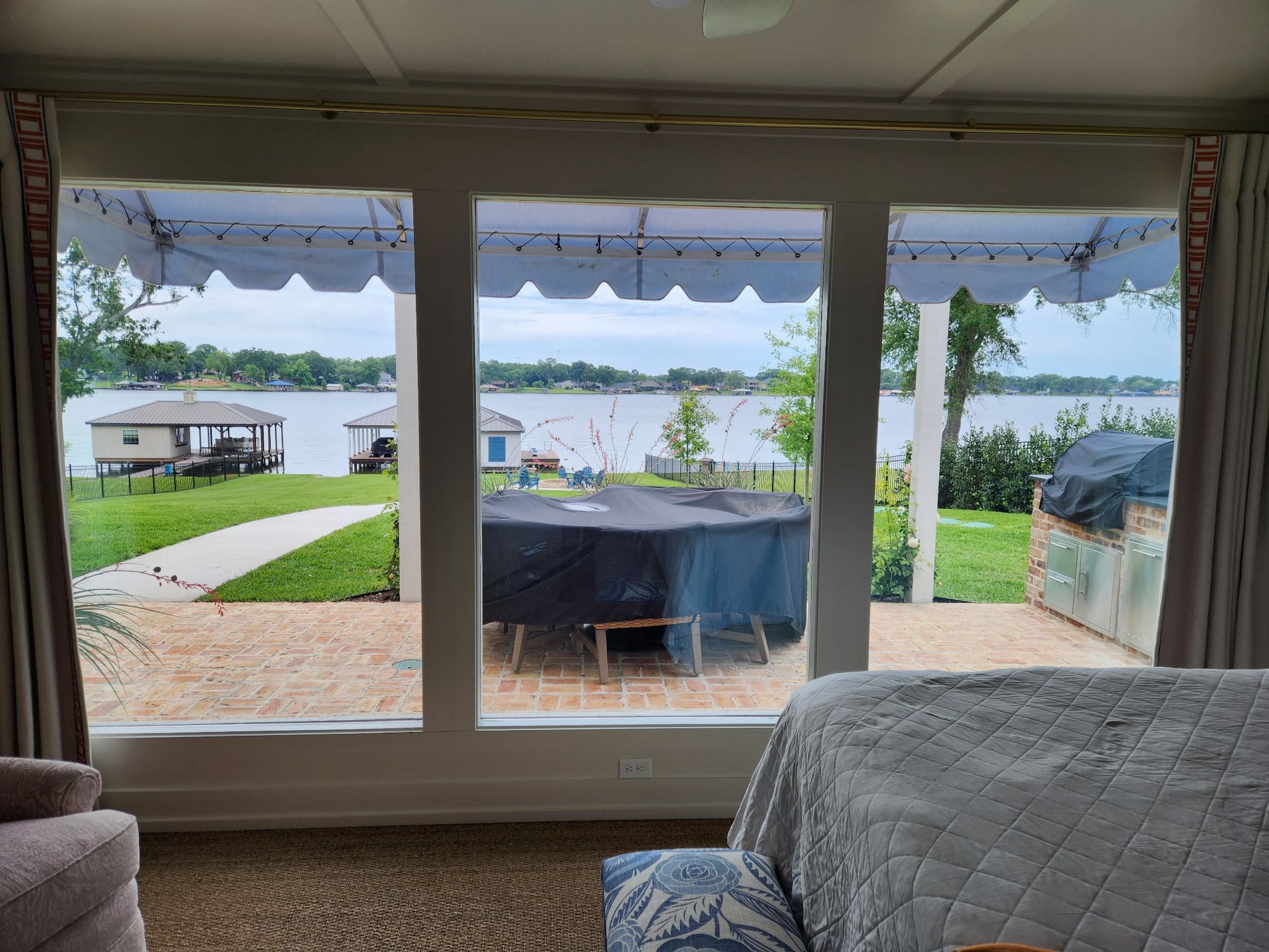 View through large windows of a lakeside yard with a dock and patio, seen from a cozy bedroom.