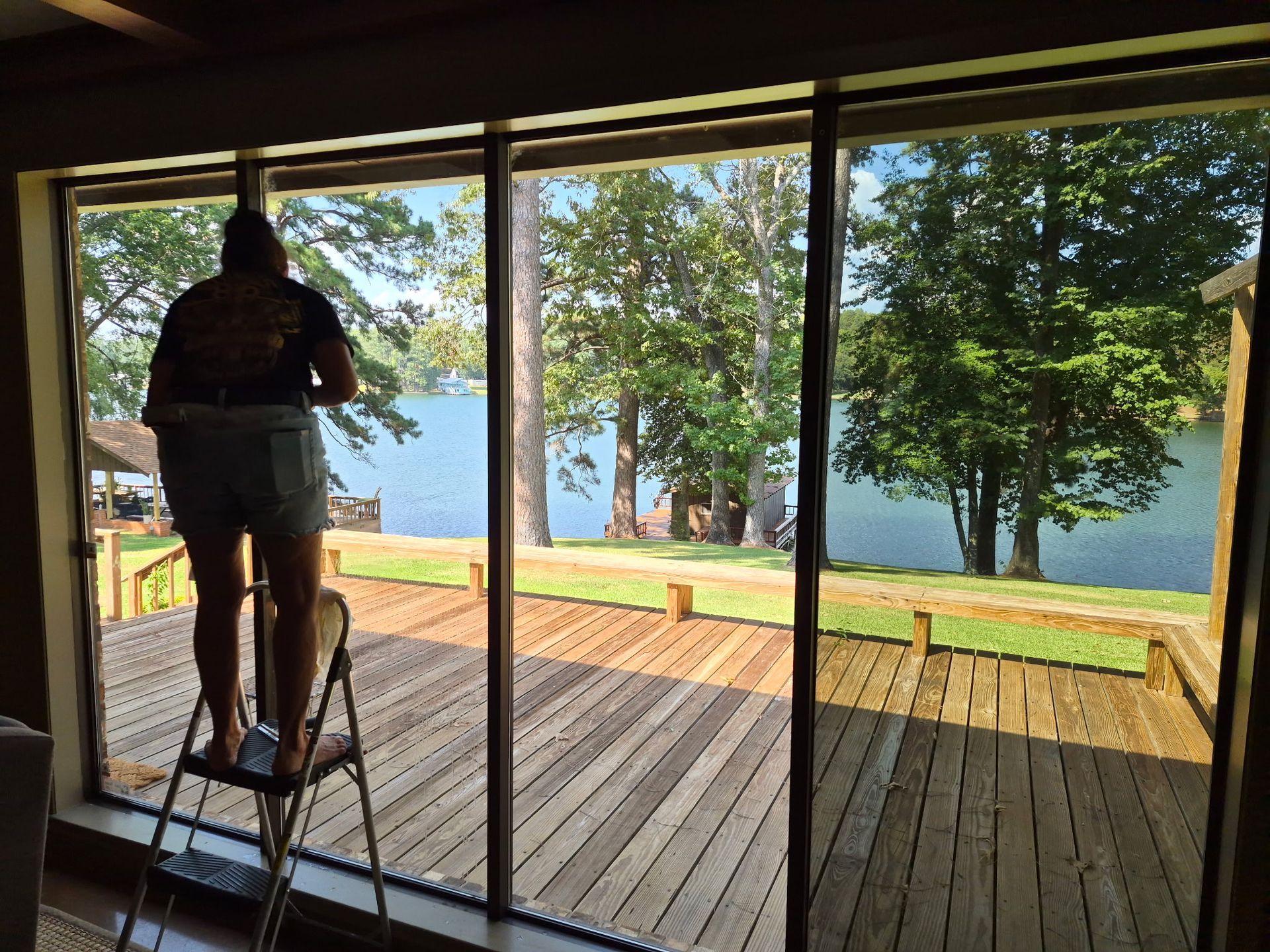 Person mowing a wooden deck beside a lake, seen through large sliding glass doors.