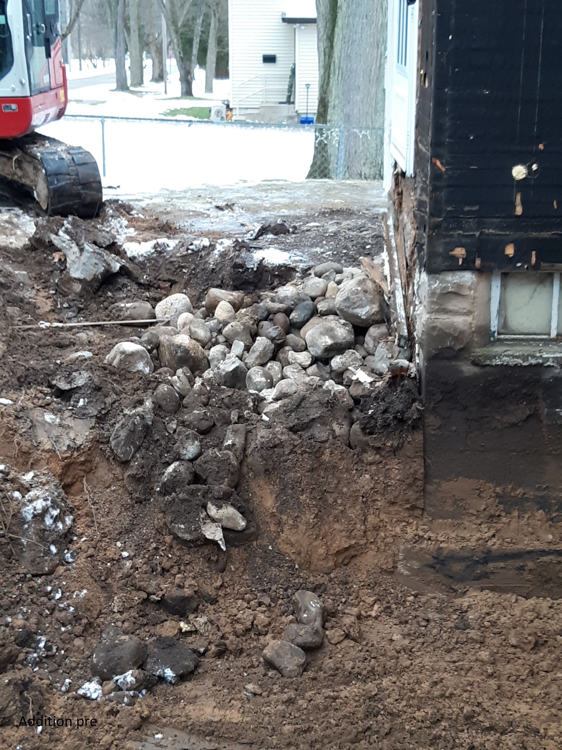 Excavator digging a muddy trench beside a building, with a pile of rocks and soil in the foreground