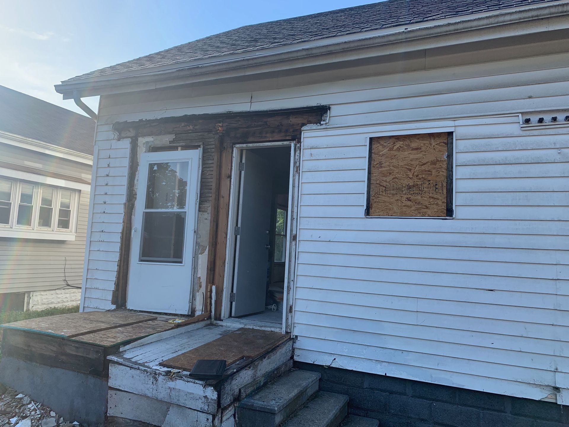 Small house exterior with damaged front steps and an open front door, boarded-up window, and white siding.
