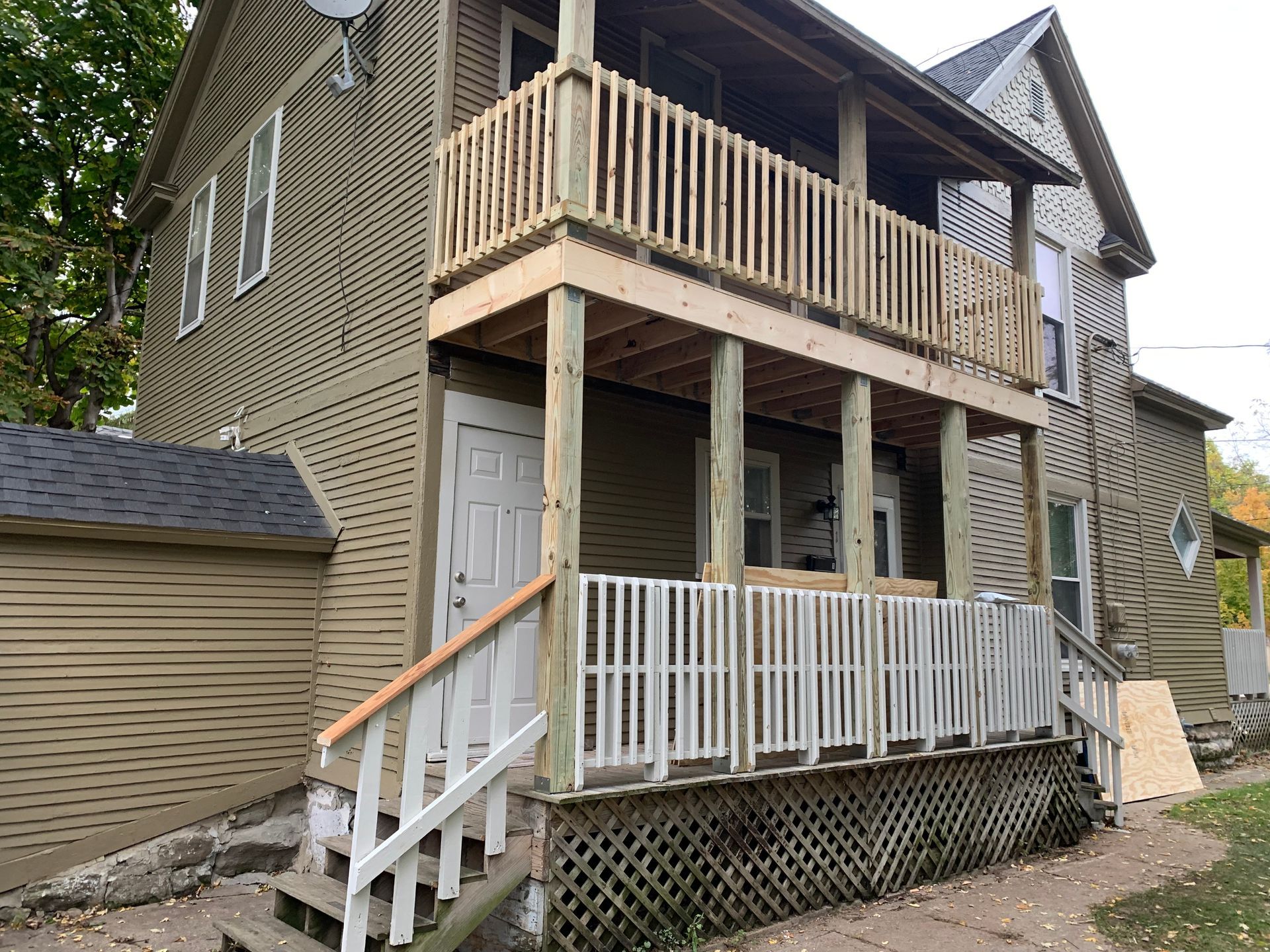 Two-story tan house with a wooden balcony, front porch, and white railing, viewed from the side