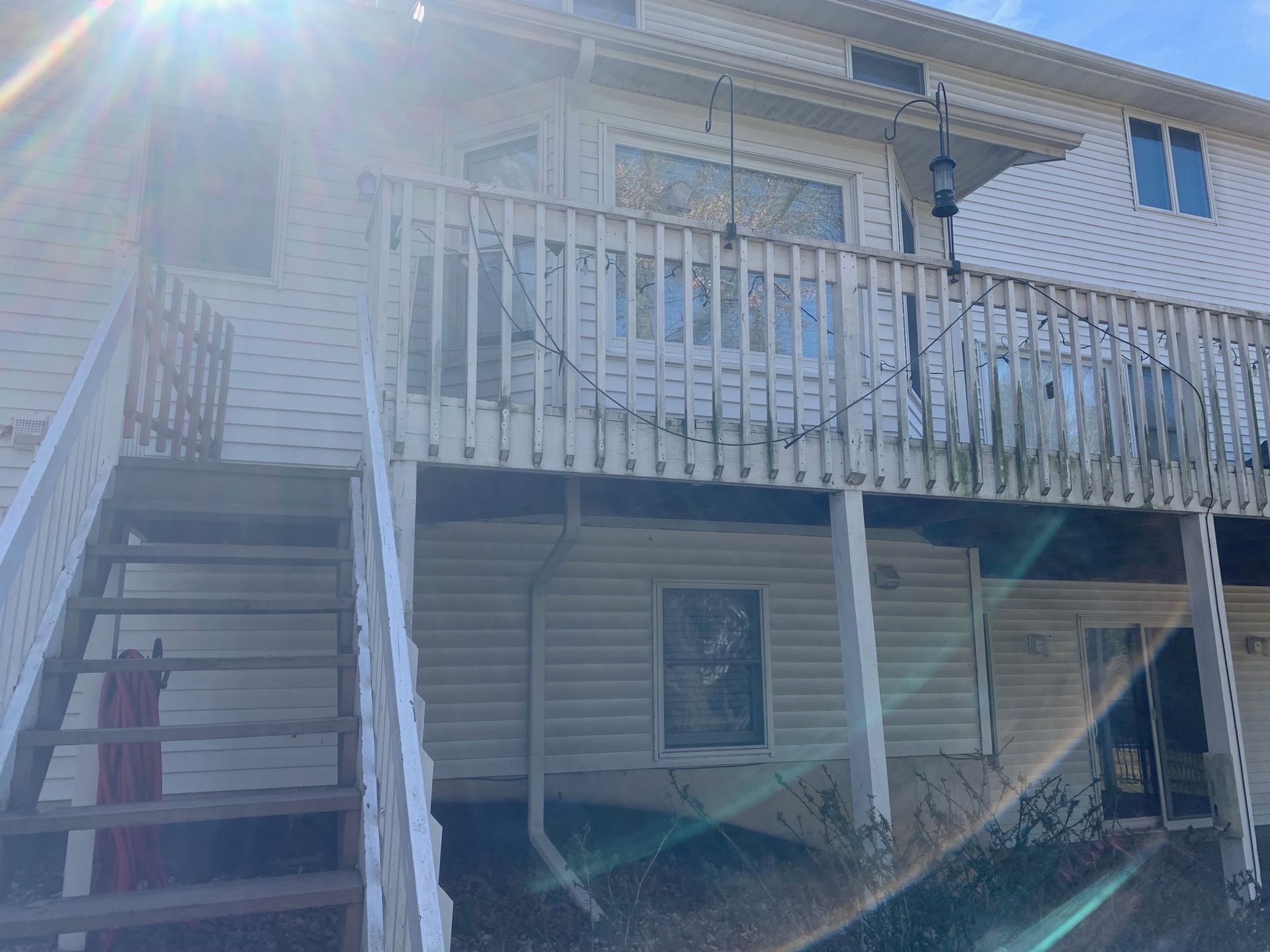 Two-story house with white siding, an exterior staircase, and a weathered wooden balcony in bright sunlight