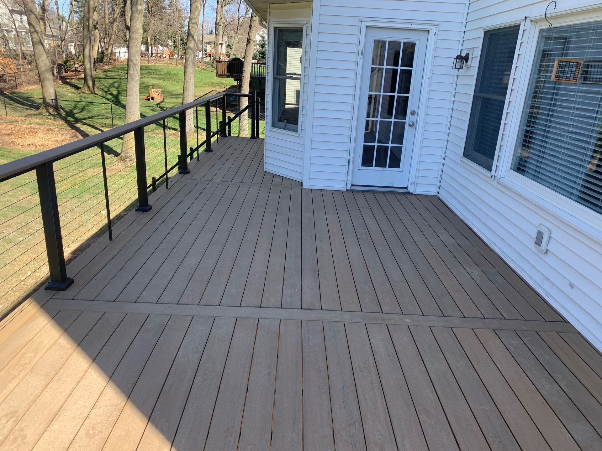 Wooden deck with black railing beside a white house, overlooking a wooded yard