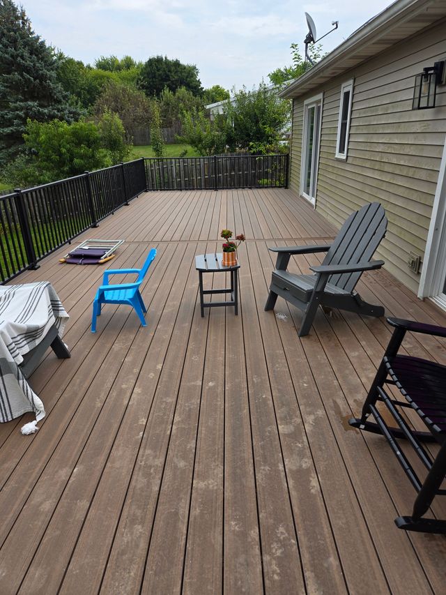 Wooden deck with scattered lawn chairs and a small table beside a house, overlooking trees and a fence.
