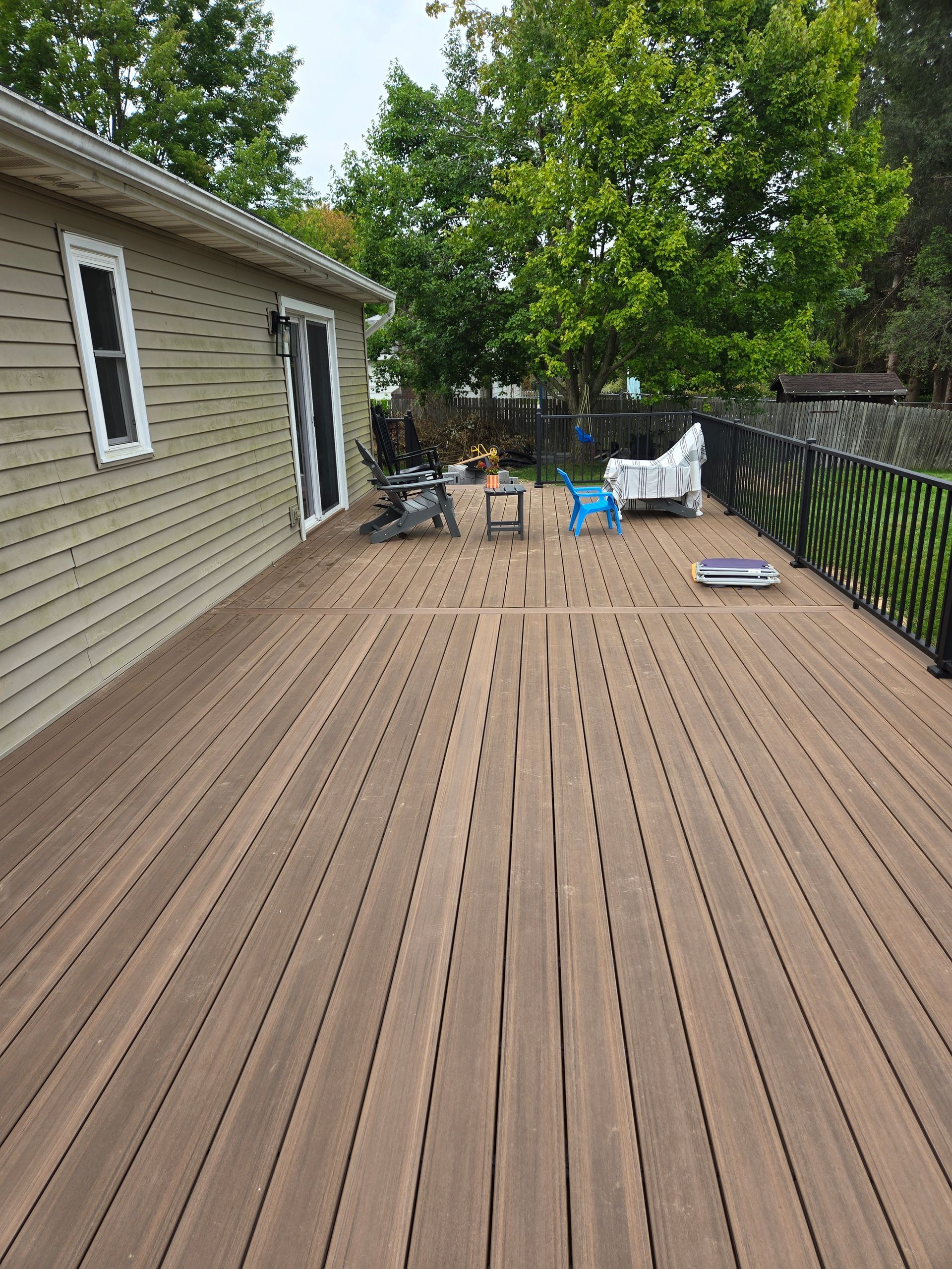Wooden backyard deck beside a house, with patio chairs, tools, and trees along the fence.