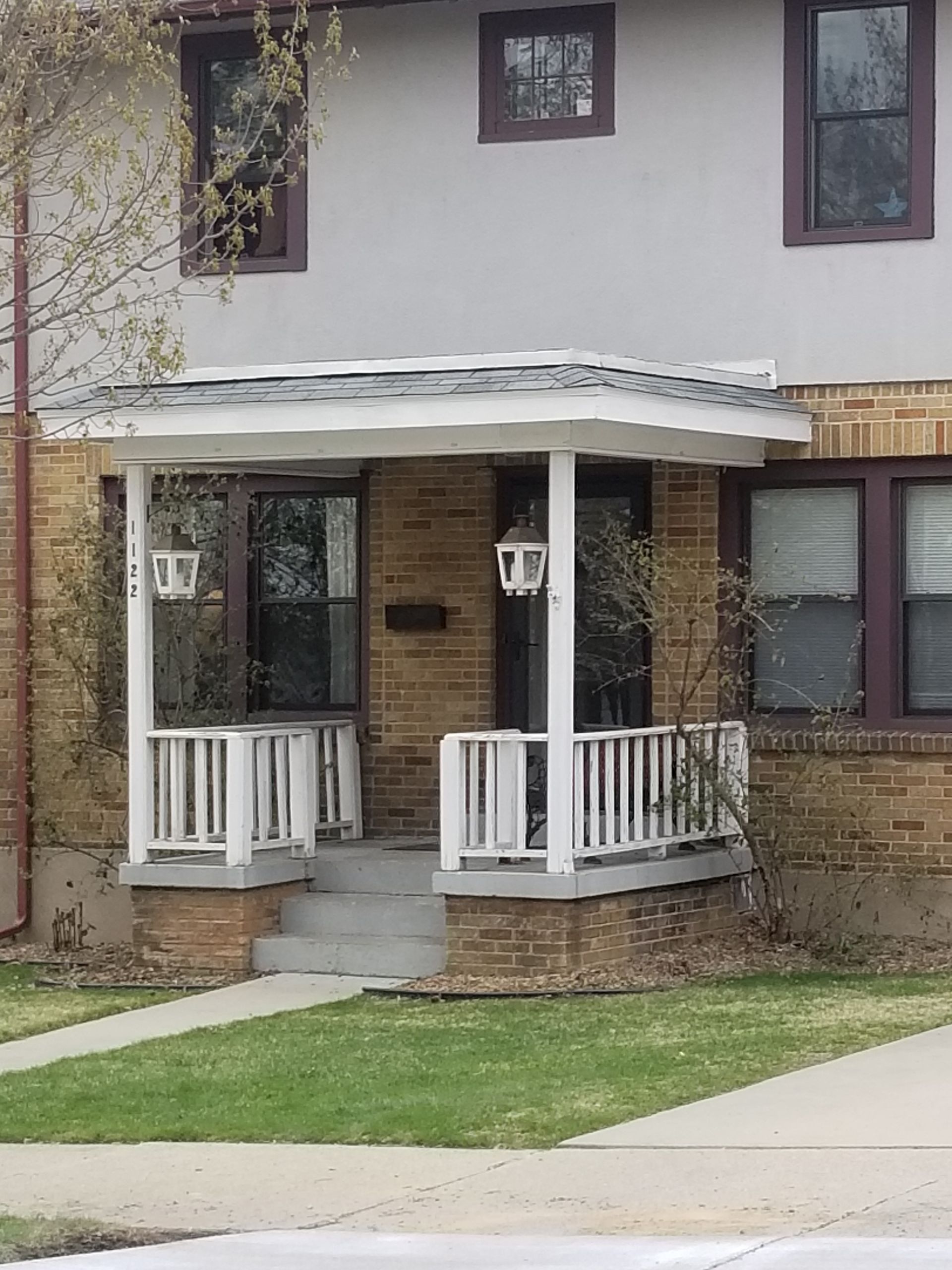 Front porch of a beige townhouse with white railings, brown brick accents, and a small lawn in front