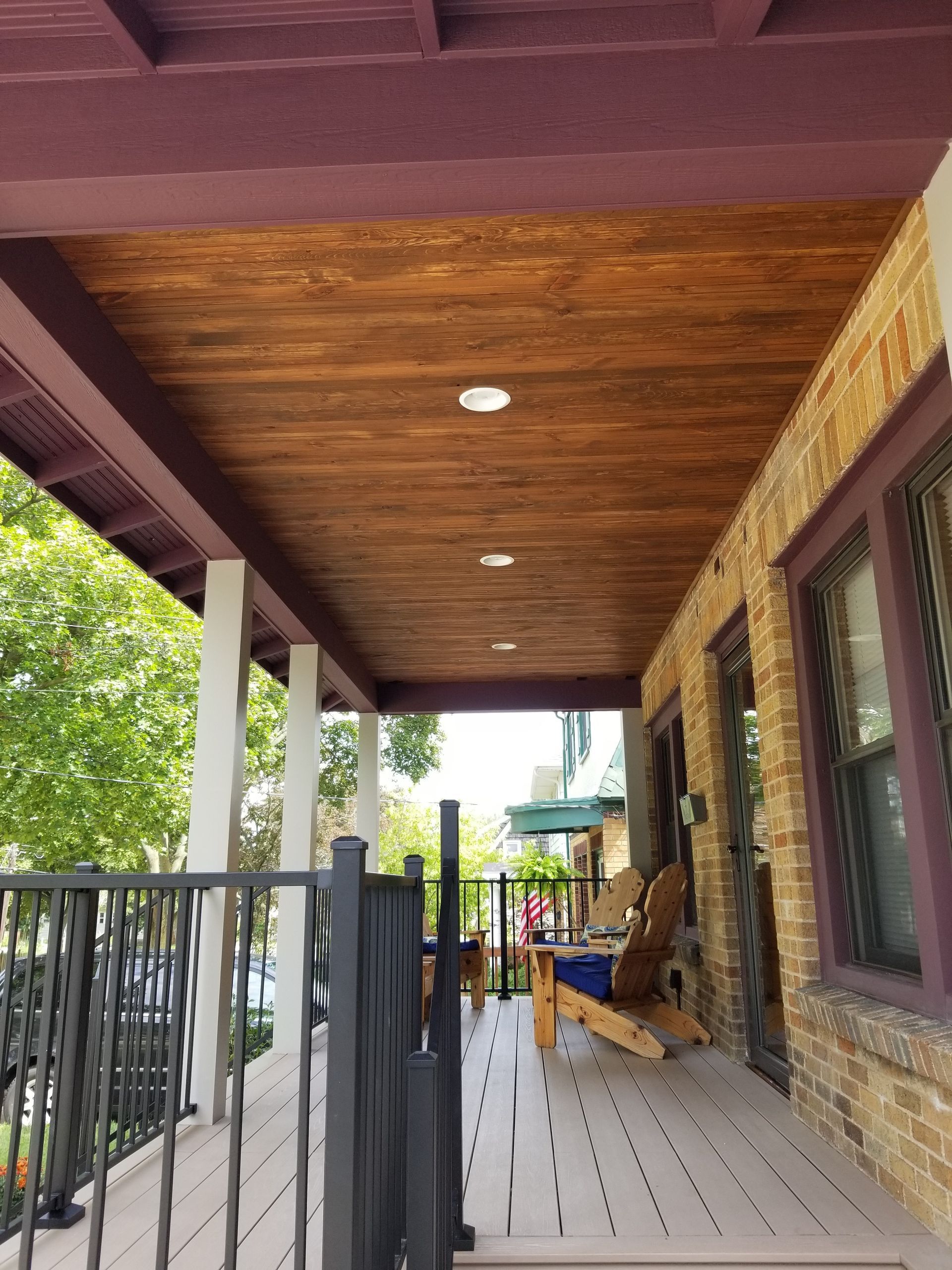 Covered porch with wood ceiling, stone wall, black railing, and potted chairs along a white deck.
