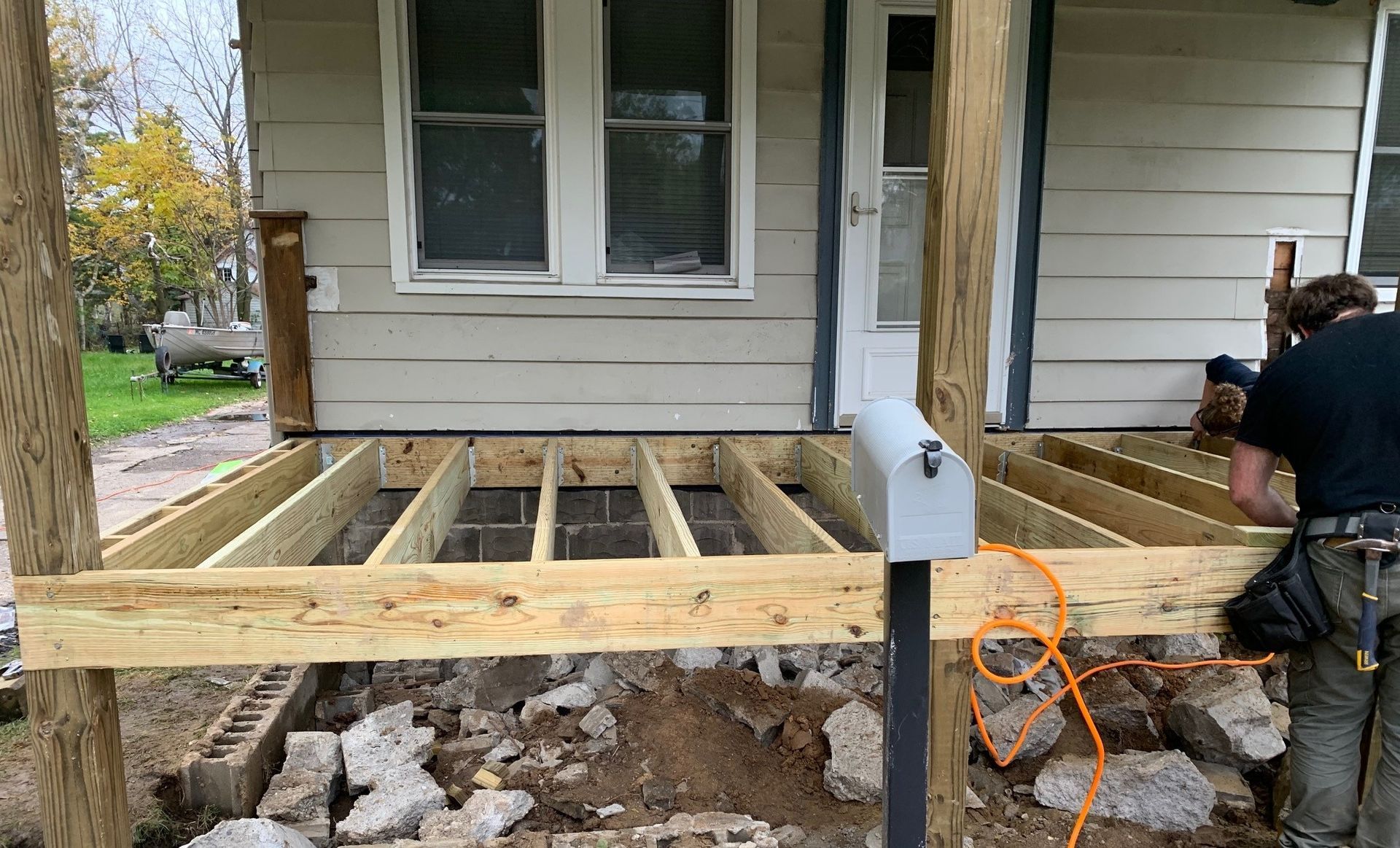 House porch under construction with exposed floor joists, a concrete footing, and a worker using tools nearby