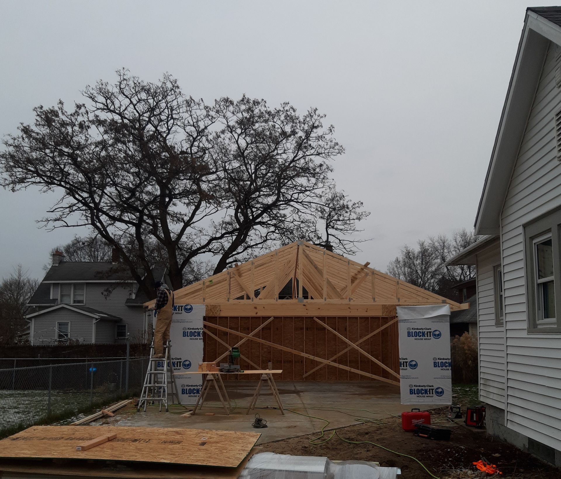 Roofing in progress on a house addition with exposed wooden trusses and construction materials in a yard.