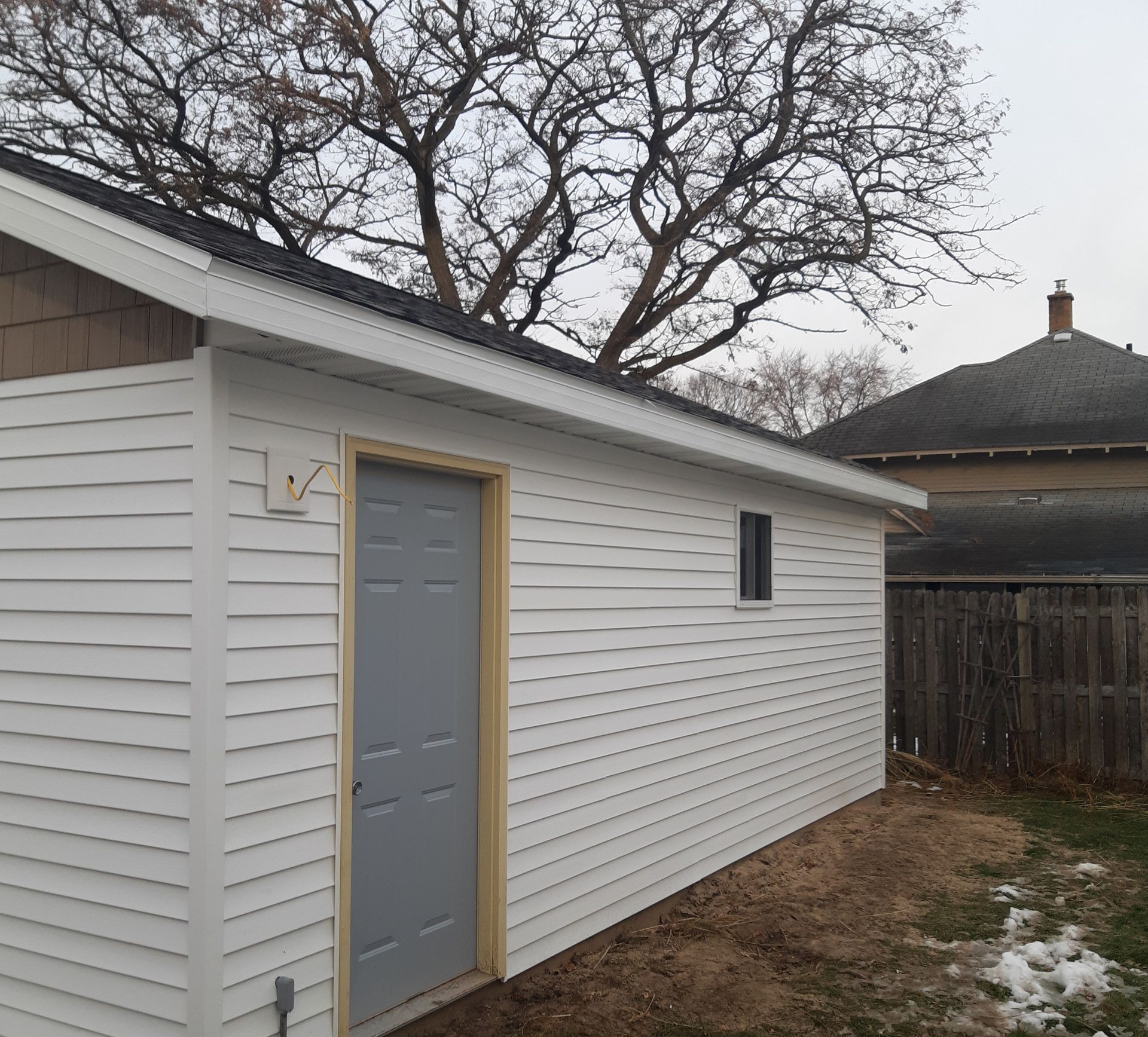 White house side with gray door, small window, and a snowy backyard under bare trees.