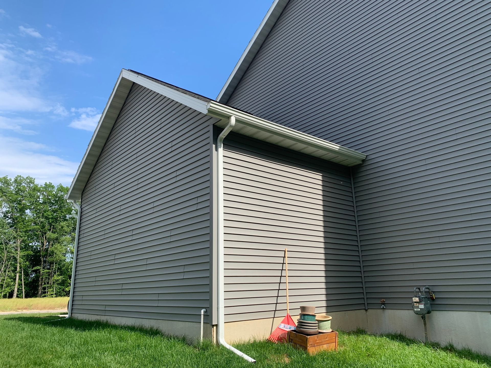 Side view of a gray house with vinyl siding, a small window well, and tools near the foundation on grass.