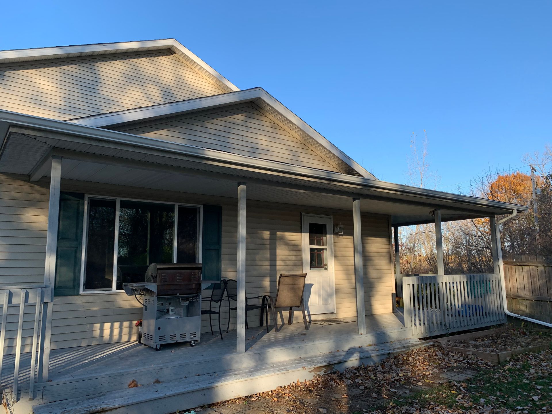 Covered porch on a beige house with grill, chairs, and white railing in autumn sunlight