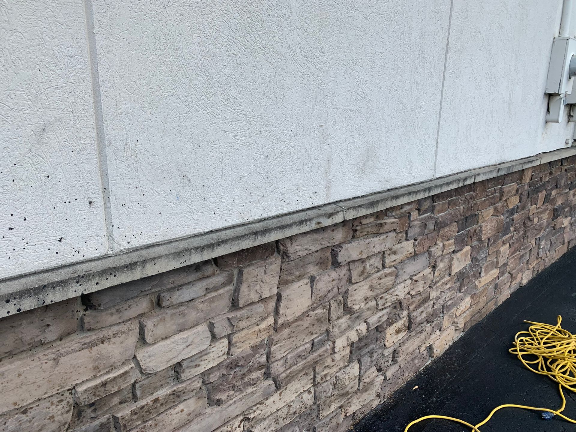 Exterior wall with light siding above a stone veneer base, beside coiled yellow cable on pavement