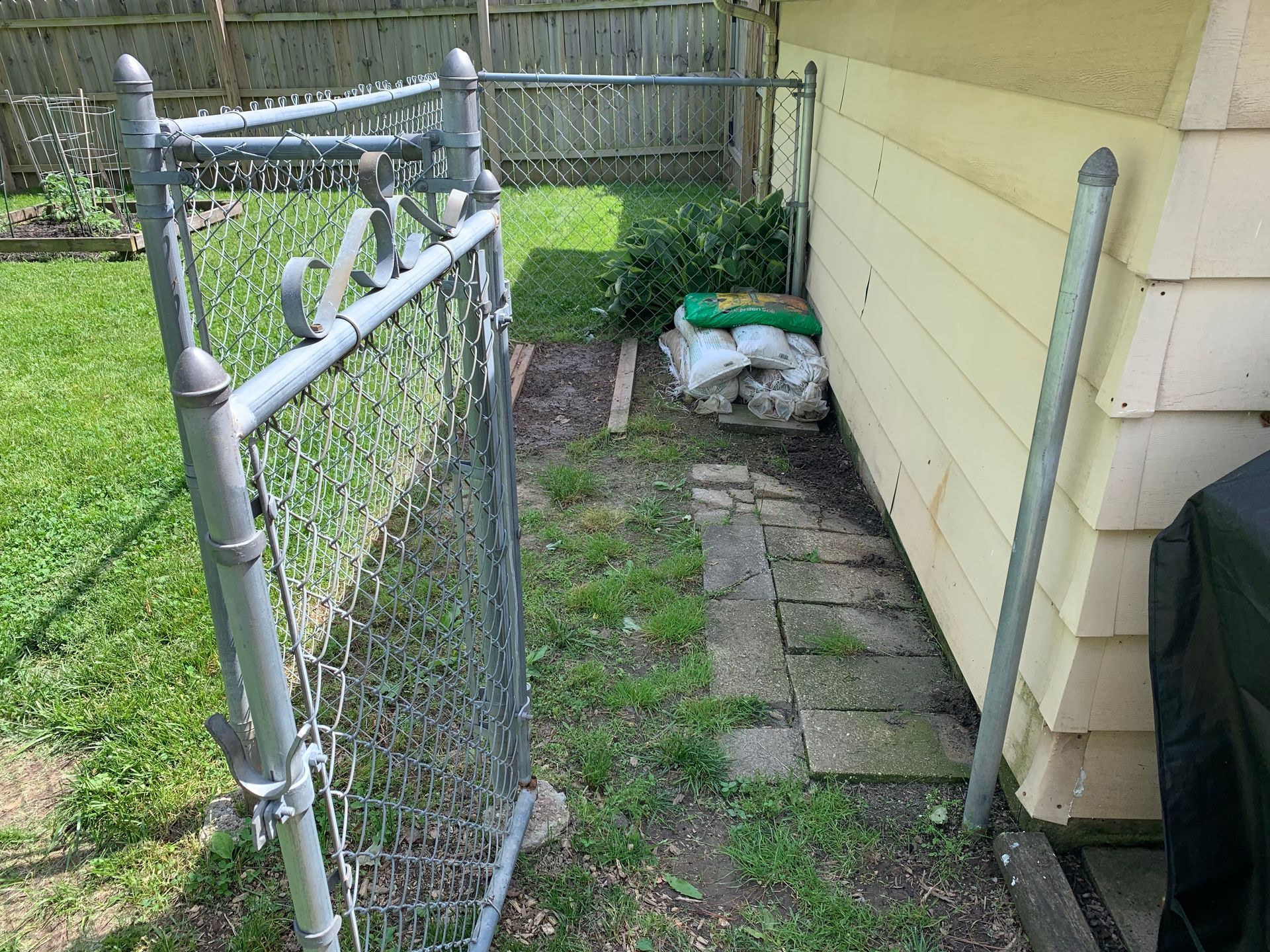 Narrow side yard with a chain-link gate, stone path, and yellow house siding, with bags near the back fence