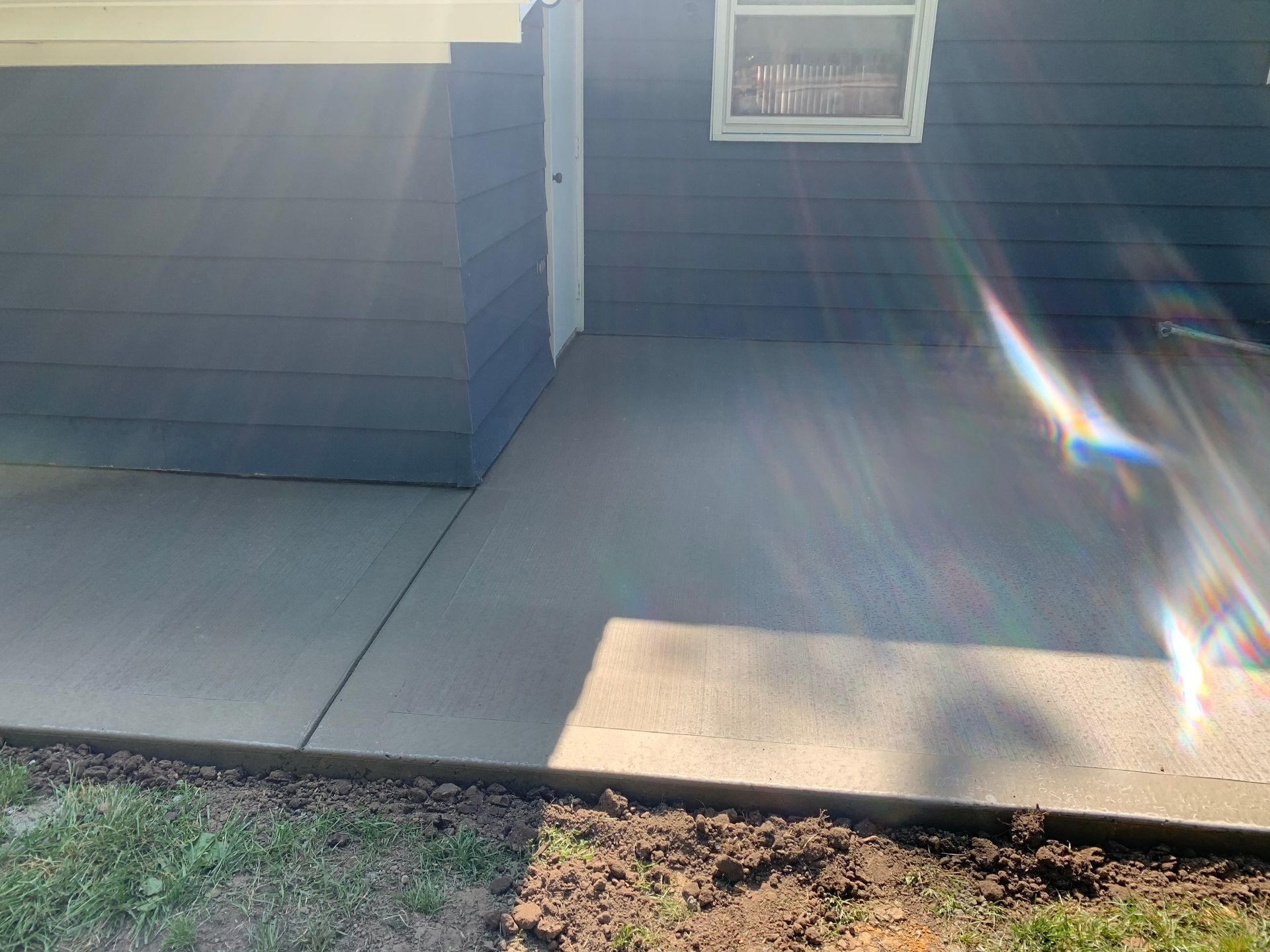 Front porch with gray siding, concrete walkway, small window, and a patch of grass in sunlight.