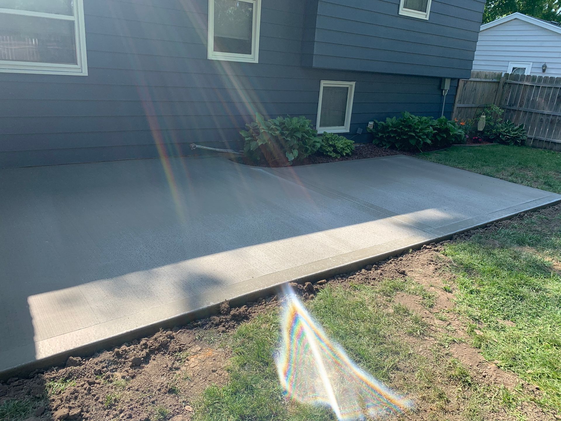 Concrete patio beside a gray house, with sun flare across the yard and grass in the foreground
