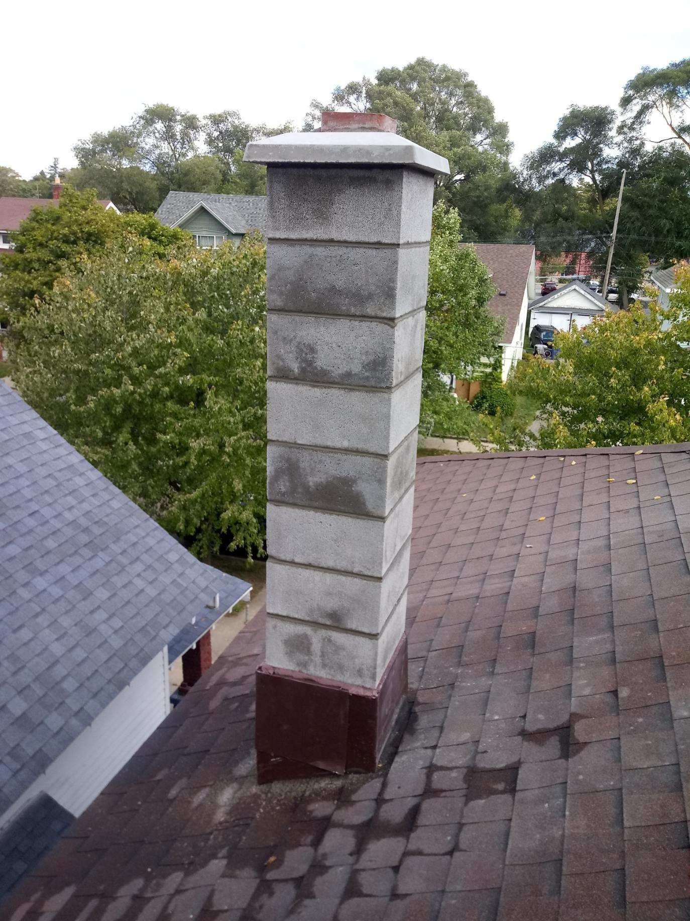 Tall gray brick chimney on a shingled rooftop with trees and houses in the background