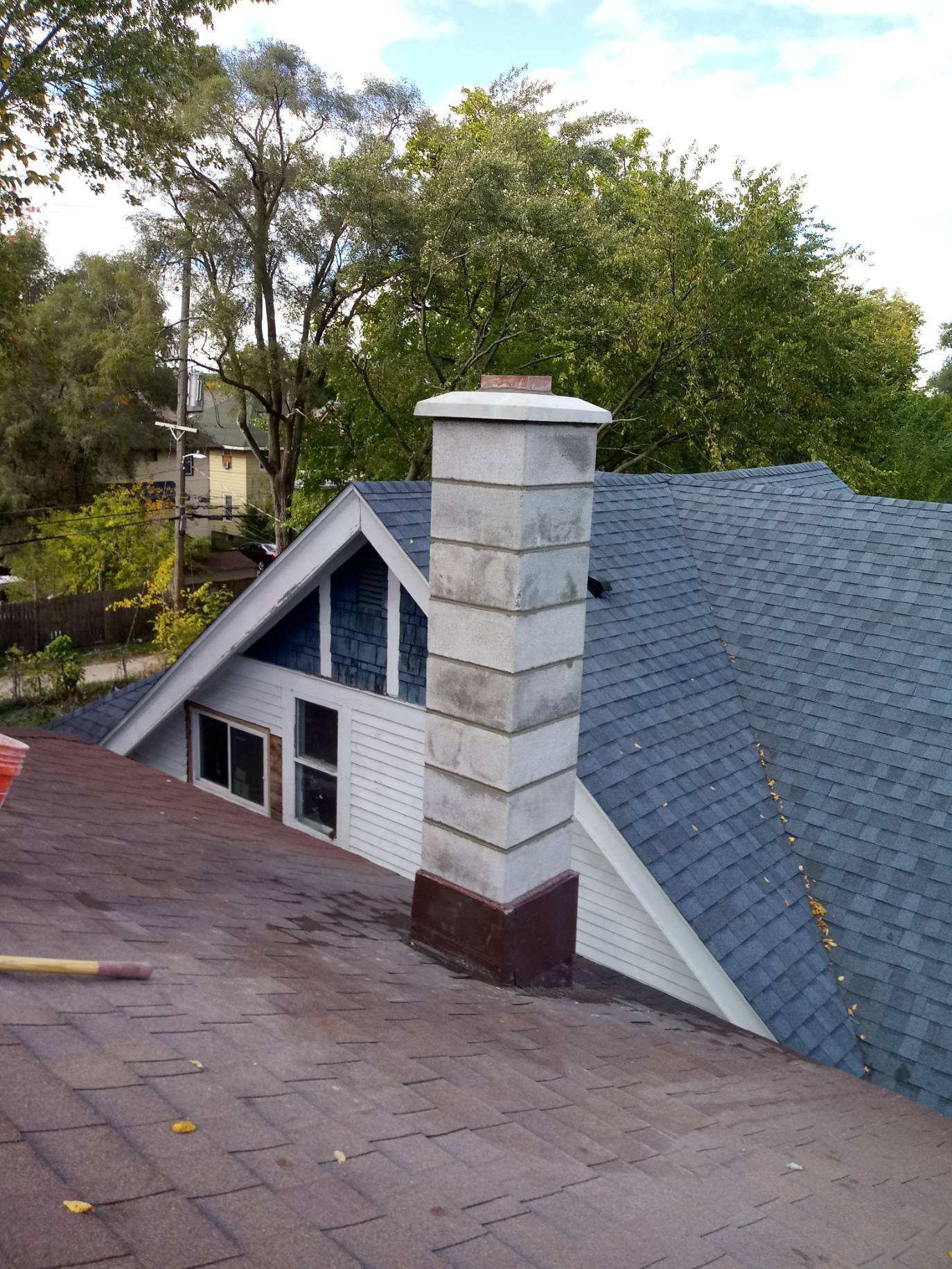 Rooftop with gray shingles and a white chimney against trees and sky