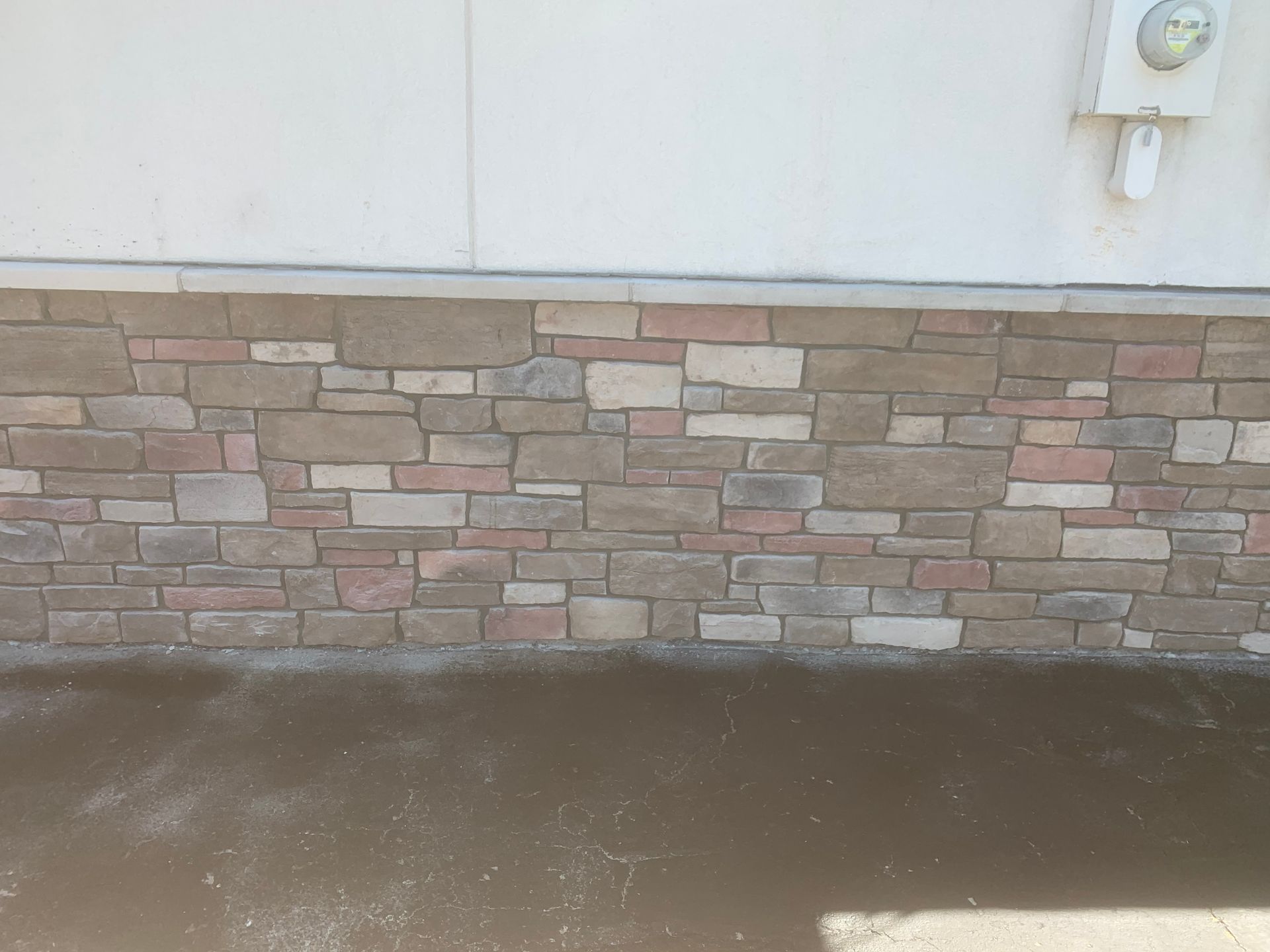 Stone veneer wall beneath a white exterior wall with a wet concrete sidewalk in front.