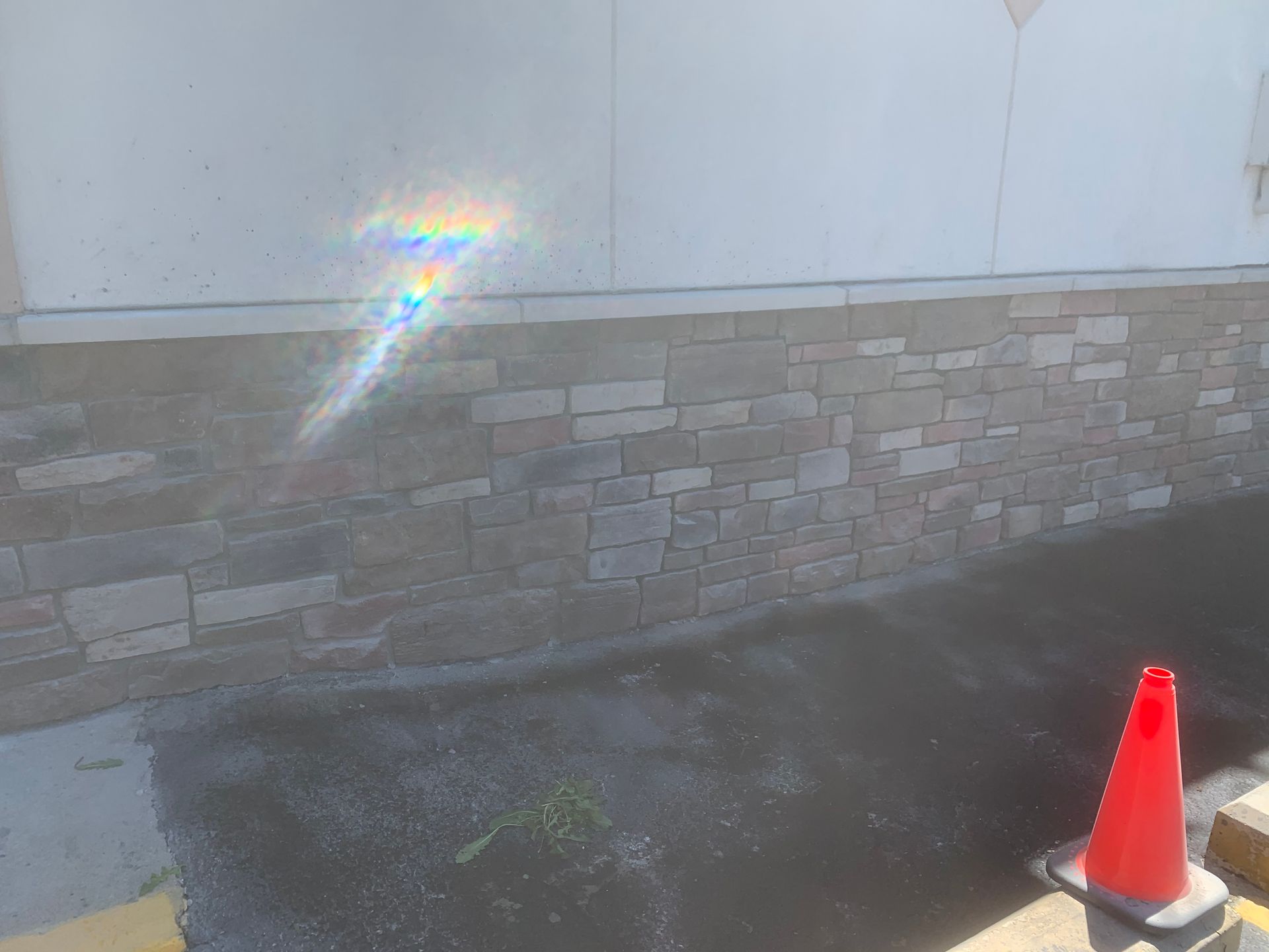 Stone wall with bright sun glare and a red traffic cone on wet pavement.