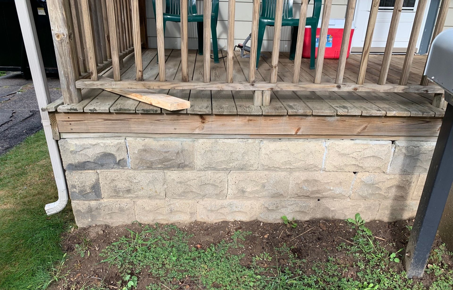 Weathered porch with wooden railing and stone foundation, showing a damaged lower wall and patchy grass below.