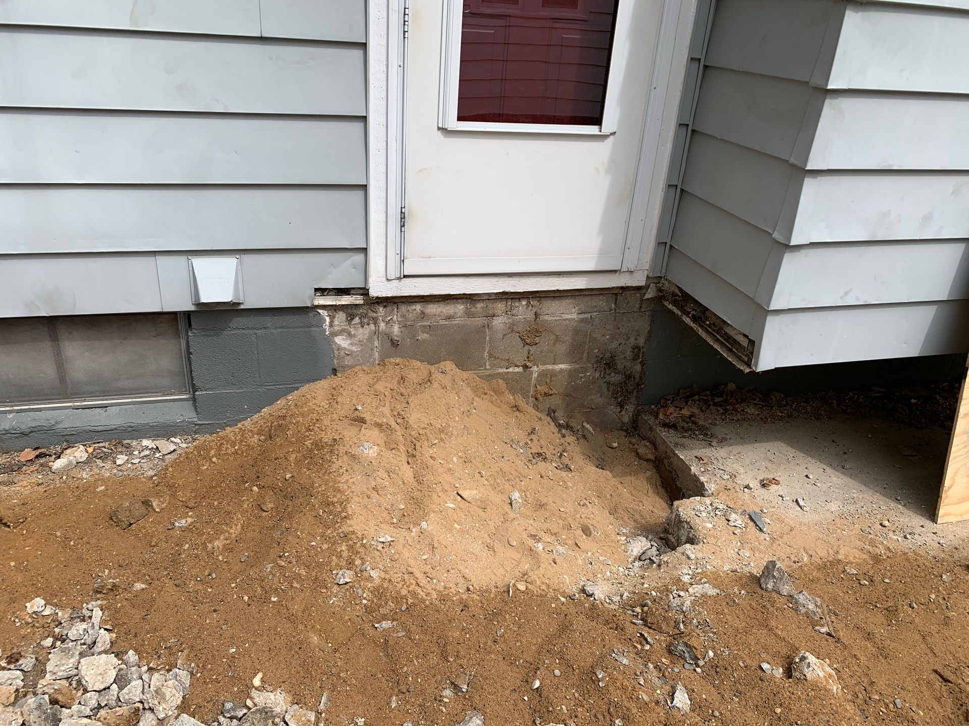 Pile of dirt and gravel against a house below a white door, with a hole in the foundation.