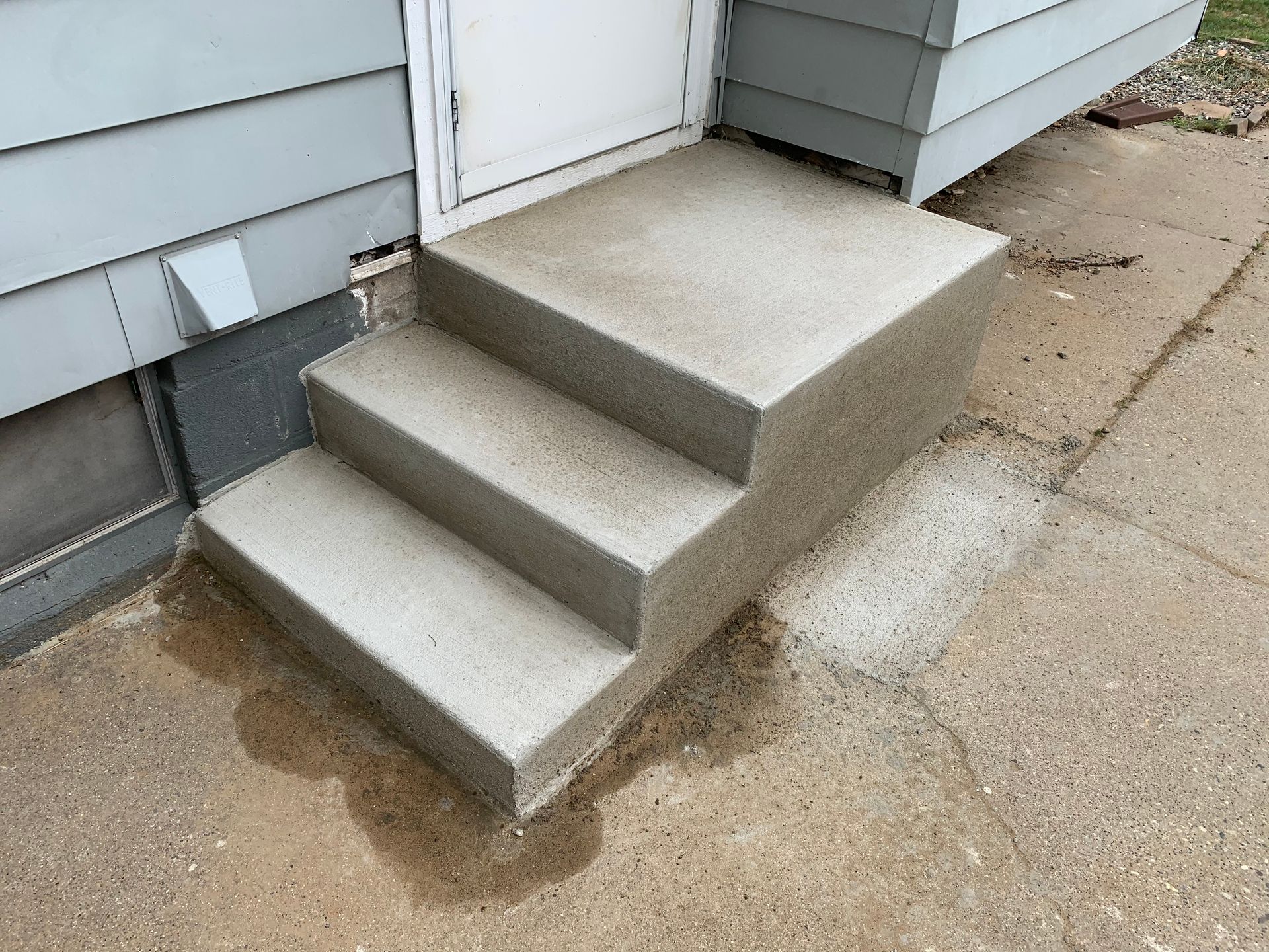 Concrete front steps leading to a white house door, with a wet driveway and light-gray siding