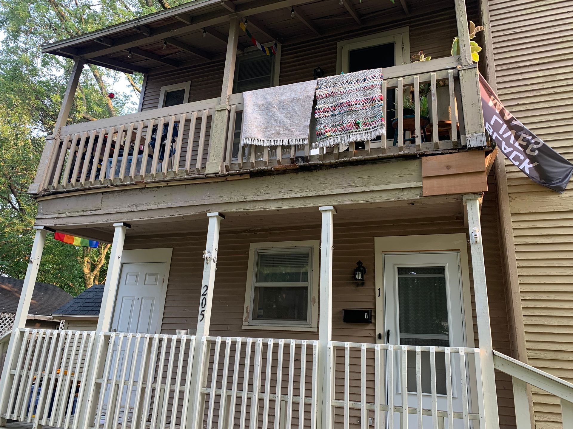Two-story wooden house with a porch and balcony, featuring white railings and a hanging banner.