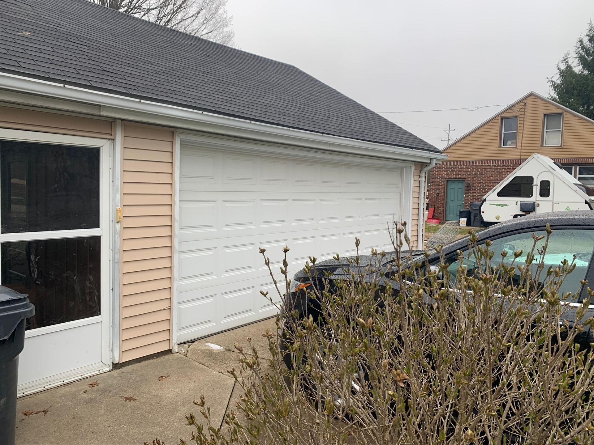 Driveway beside a beige garage with white door, overgrown shrubs in front, and parked cars nearby