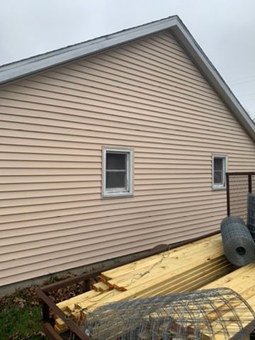 Tan siding on a house with windows; construction materials and debris piled along the foundation.
