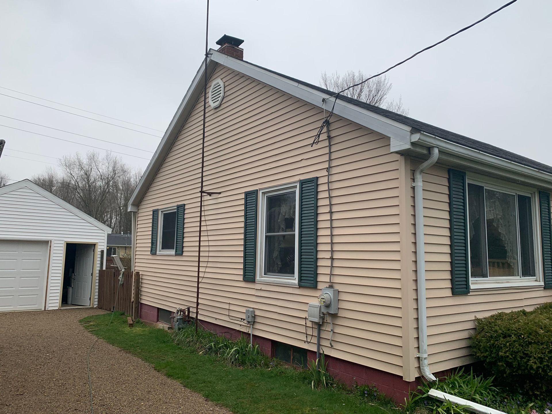 Beige vinyl-sided house with green shutters and detached garage on a cloudy day