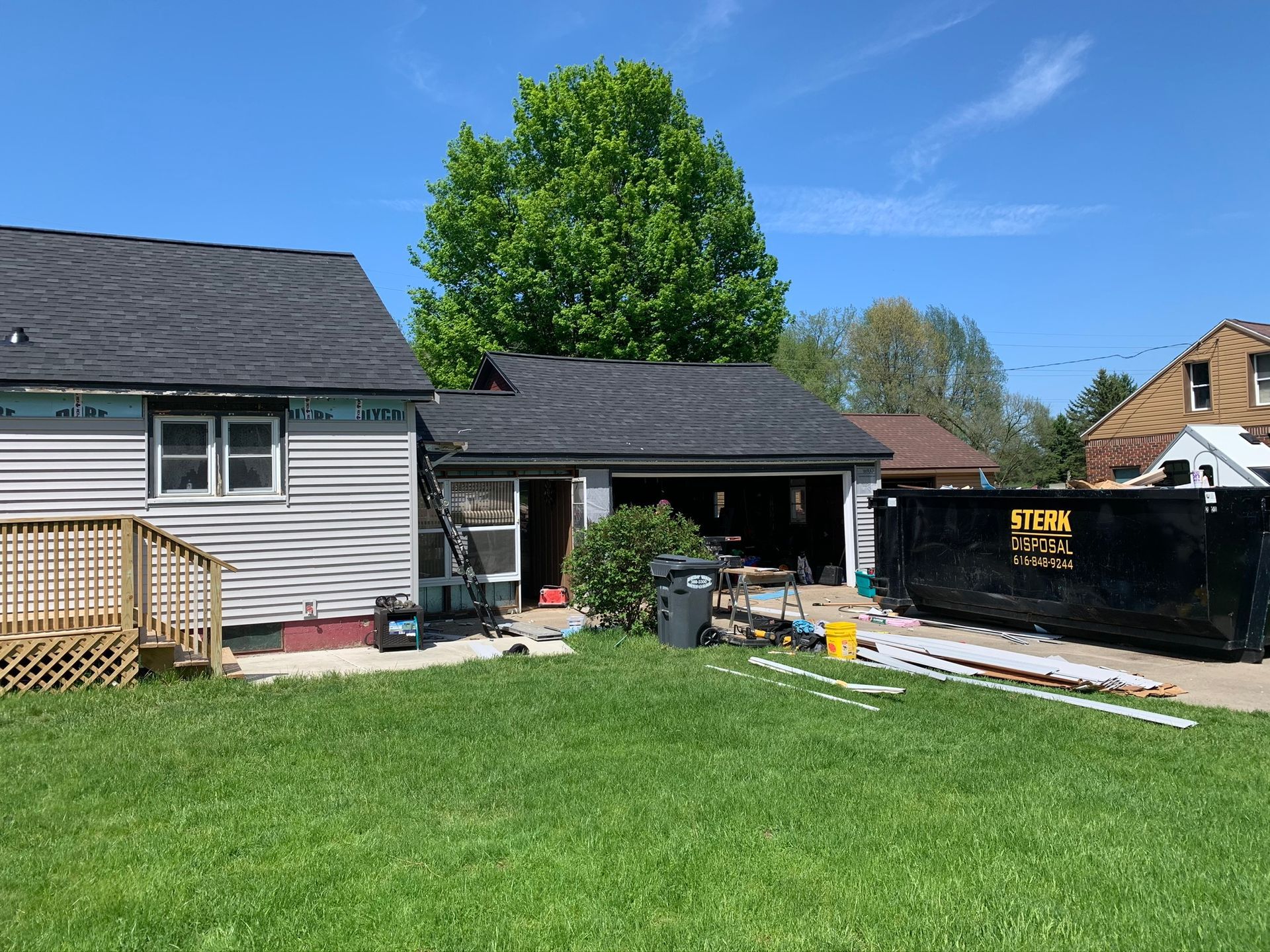 Backyard with green lawn, house and detached garage, a large tree, and construction materials on the right.