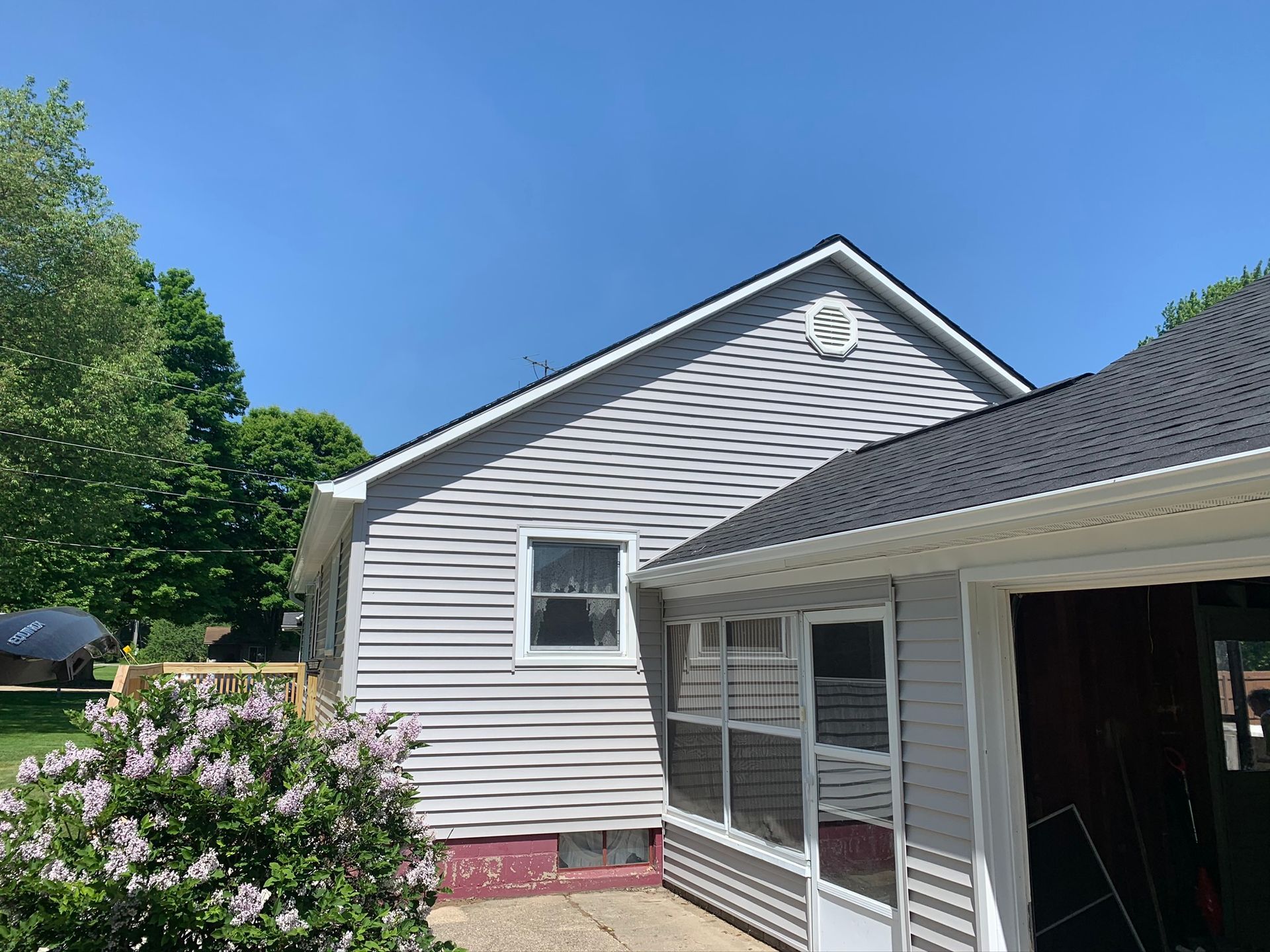 Backyard view of a gray house with screened porch, white trim, and pink flowering bushes under a clear blue sky