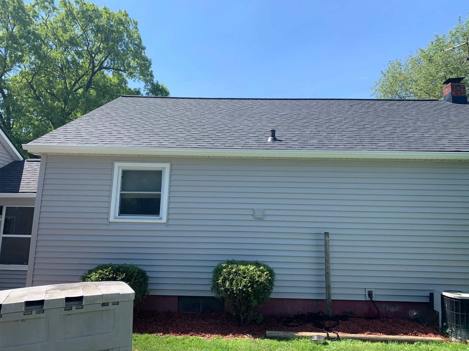 Side of a gray house with a shingled roof, small window, bushes, and a concrete block wall in front.