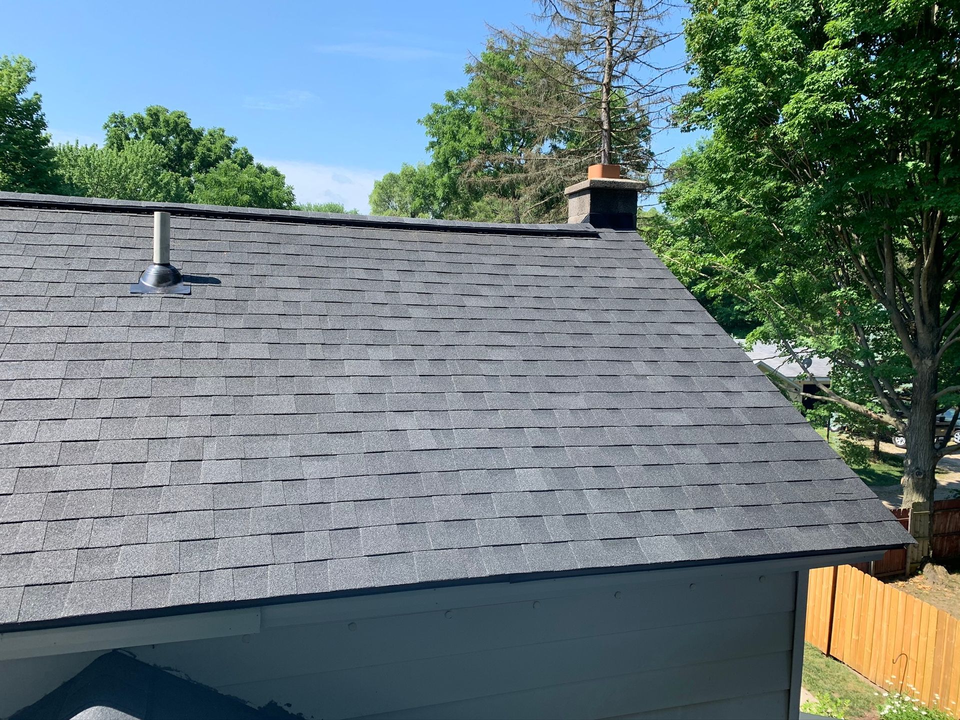 House roof with gray shingles, chimney, and trees under a blue sky