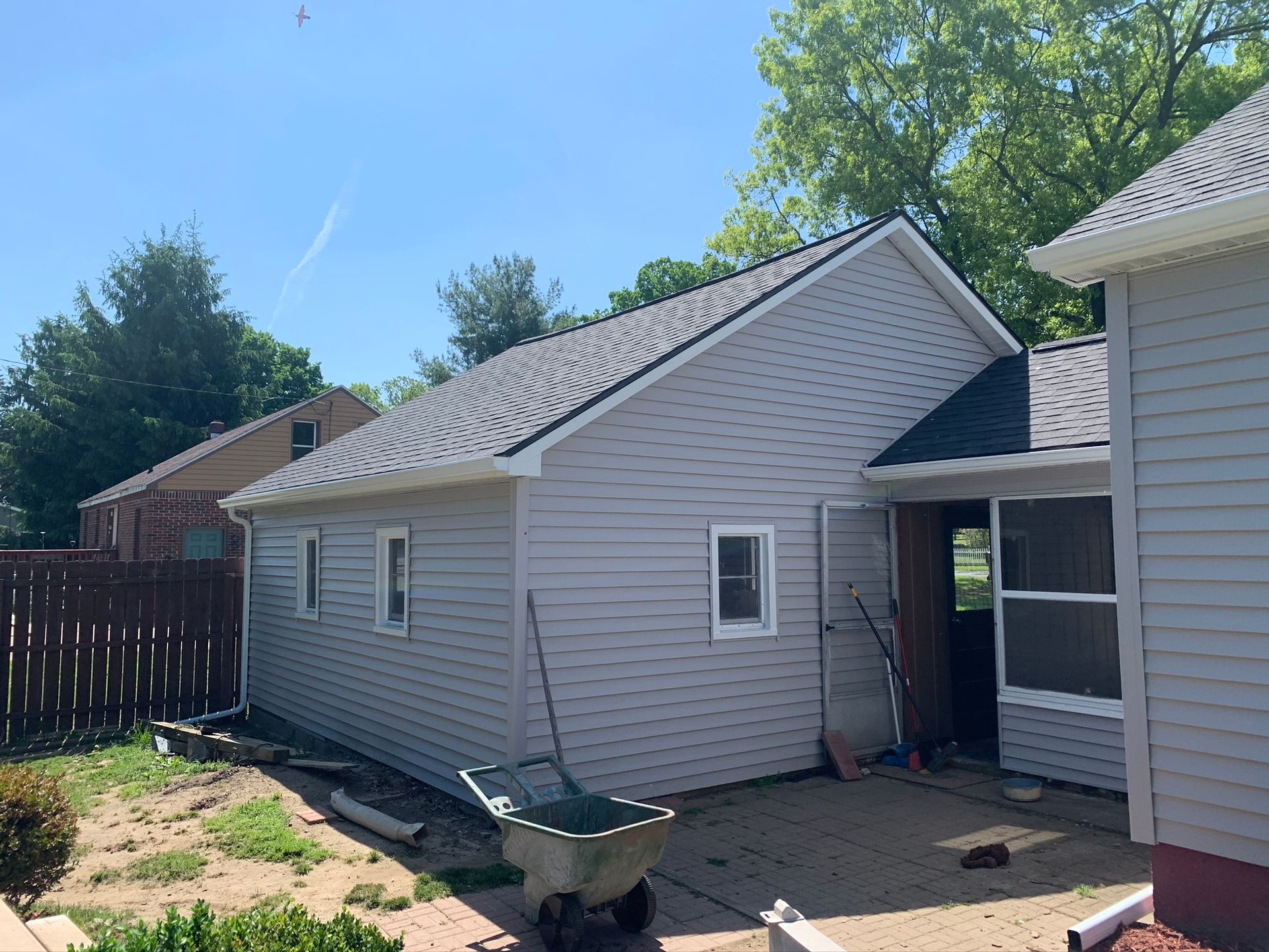 Backyard view of a gray house with a side entrance, wheelbarrow, and sunlit patio area