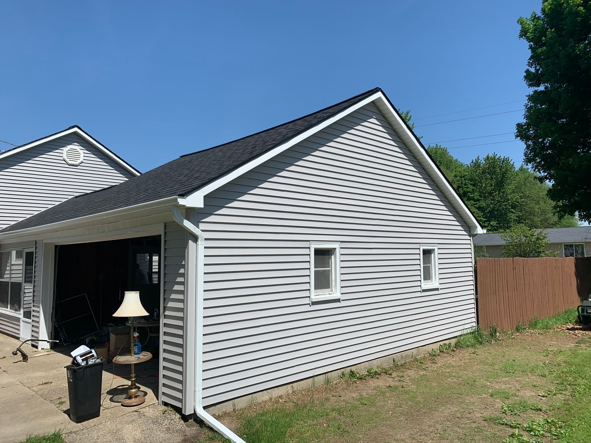 White-sided house exterior with open garage, black trash bin, and wooden fence under a blue sky