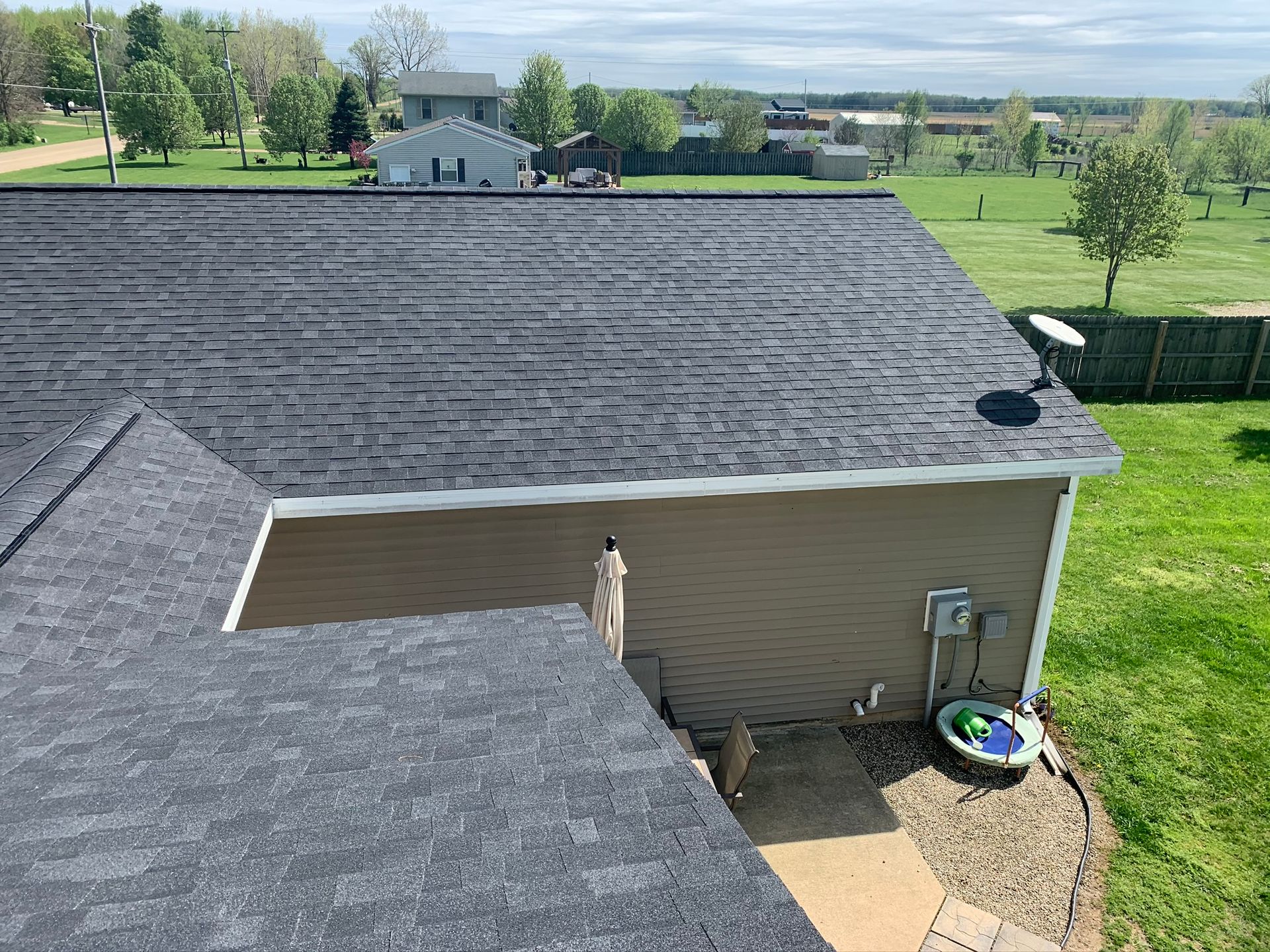 Aerial view of a house roof and backyard with green lawn, trees, and distant buildings under a clear sky