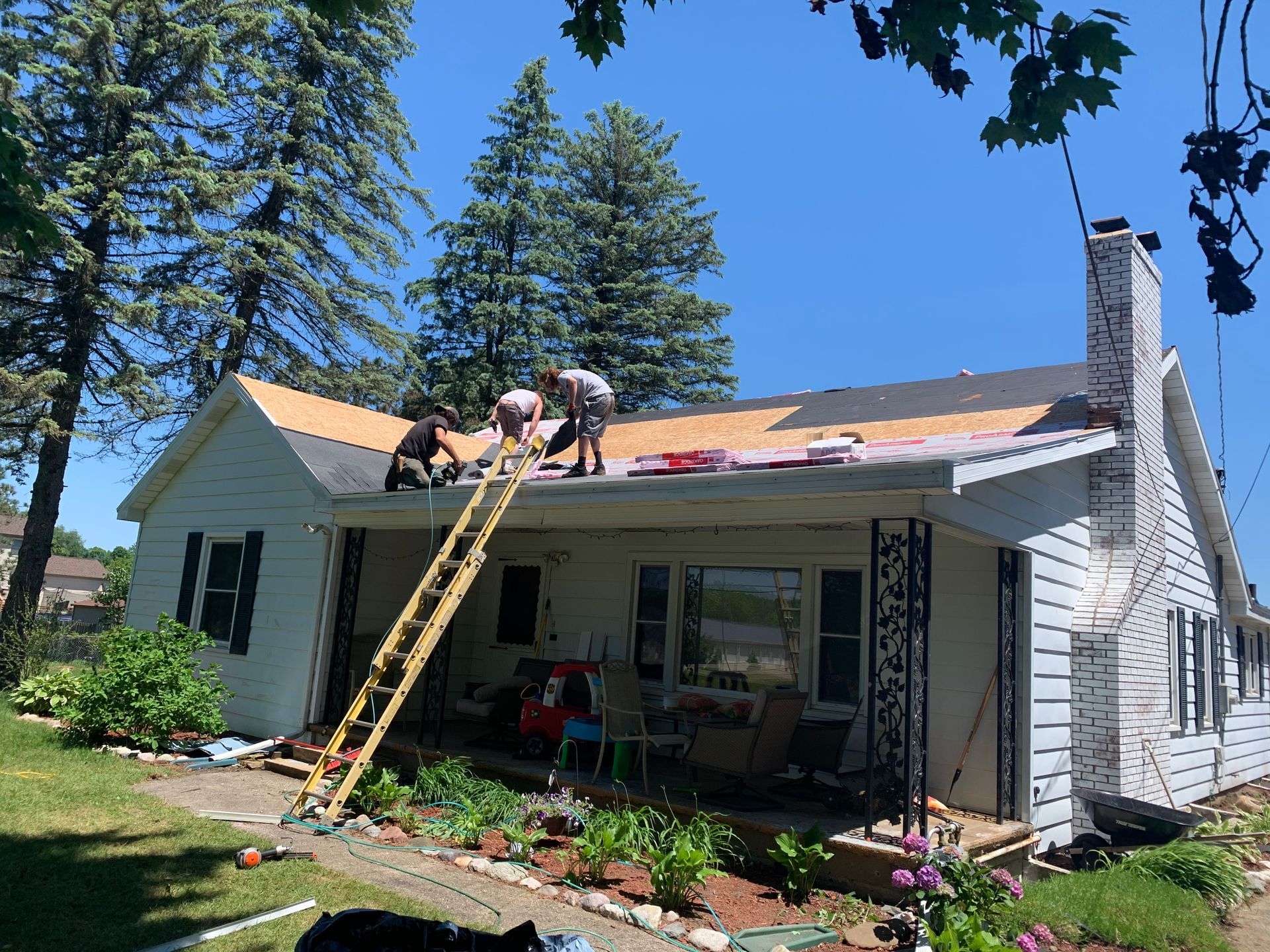 Two workers reroofing a white house with a ladder, under tall pine trees on a sunny day