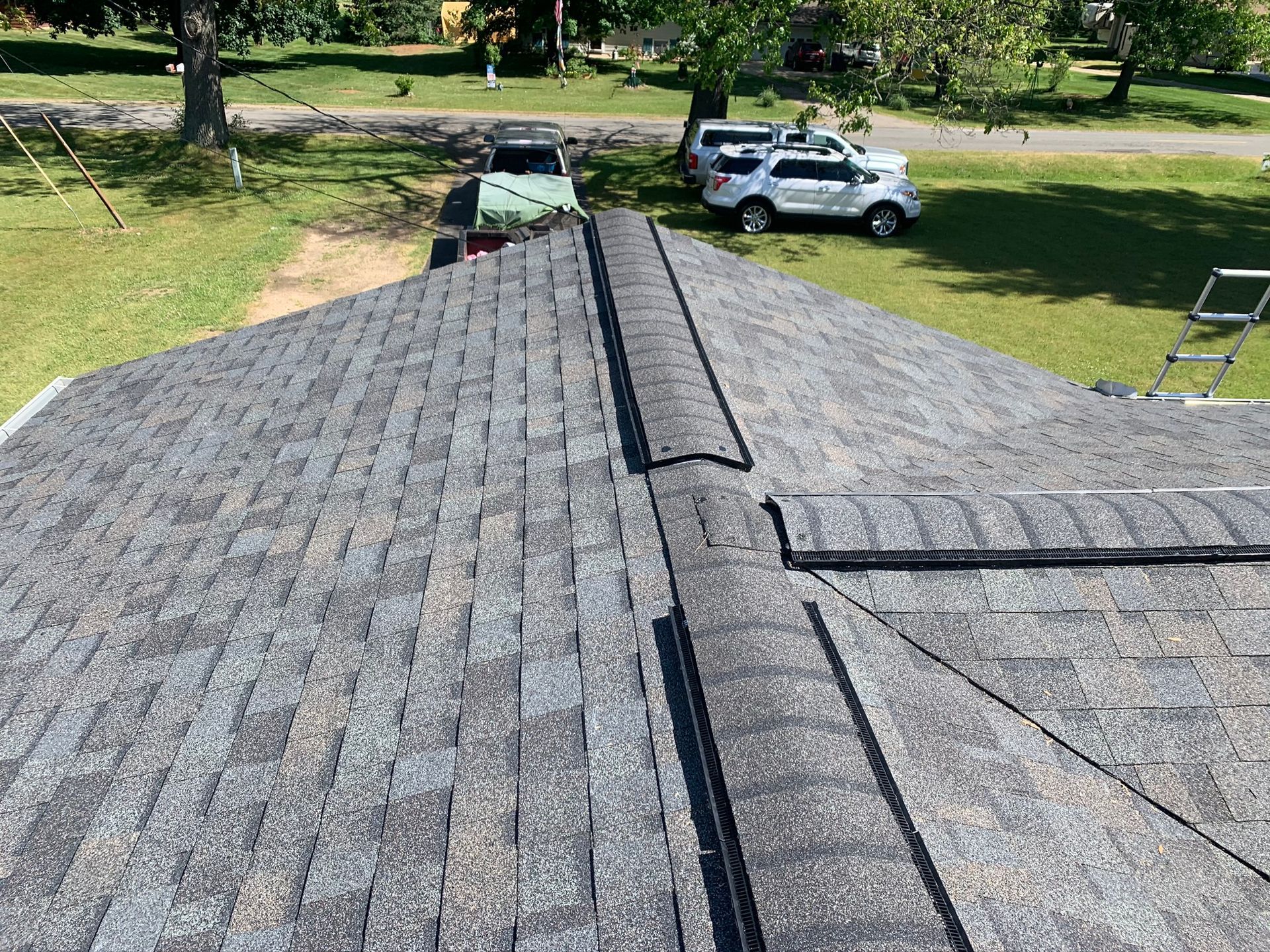 Roof shingles viewed from above with a central ridge, trees and parked vehicles in the background